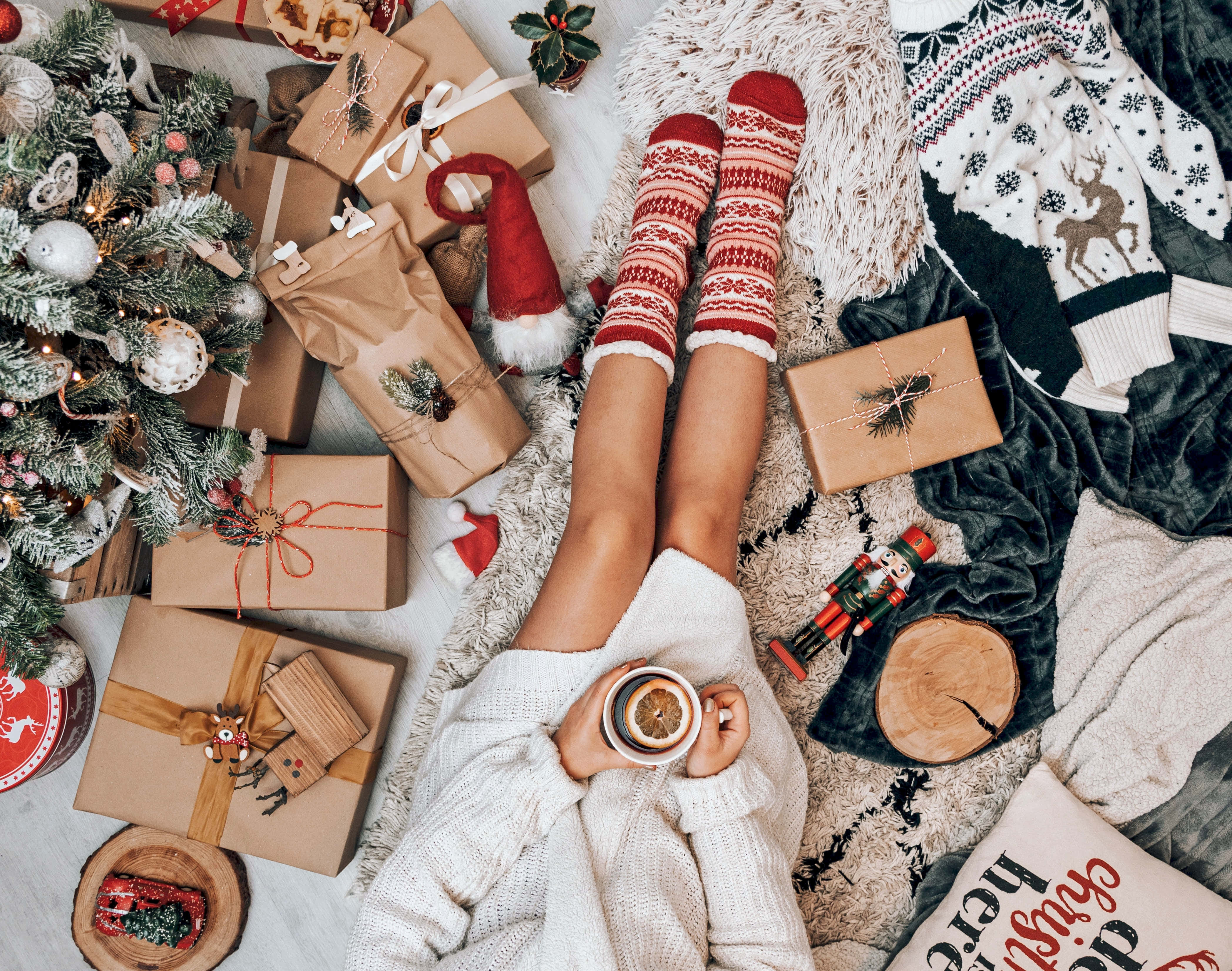 Cozy Winter Desktop 桌布 壁紙 — 俯視角度下，穿著紅色圖案襪子的舒適雙腿，手持一個被聖誕禮物和裝飾樹環繞的馬克杯。