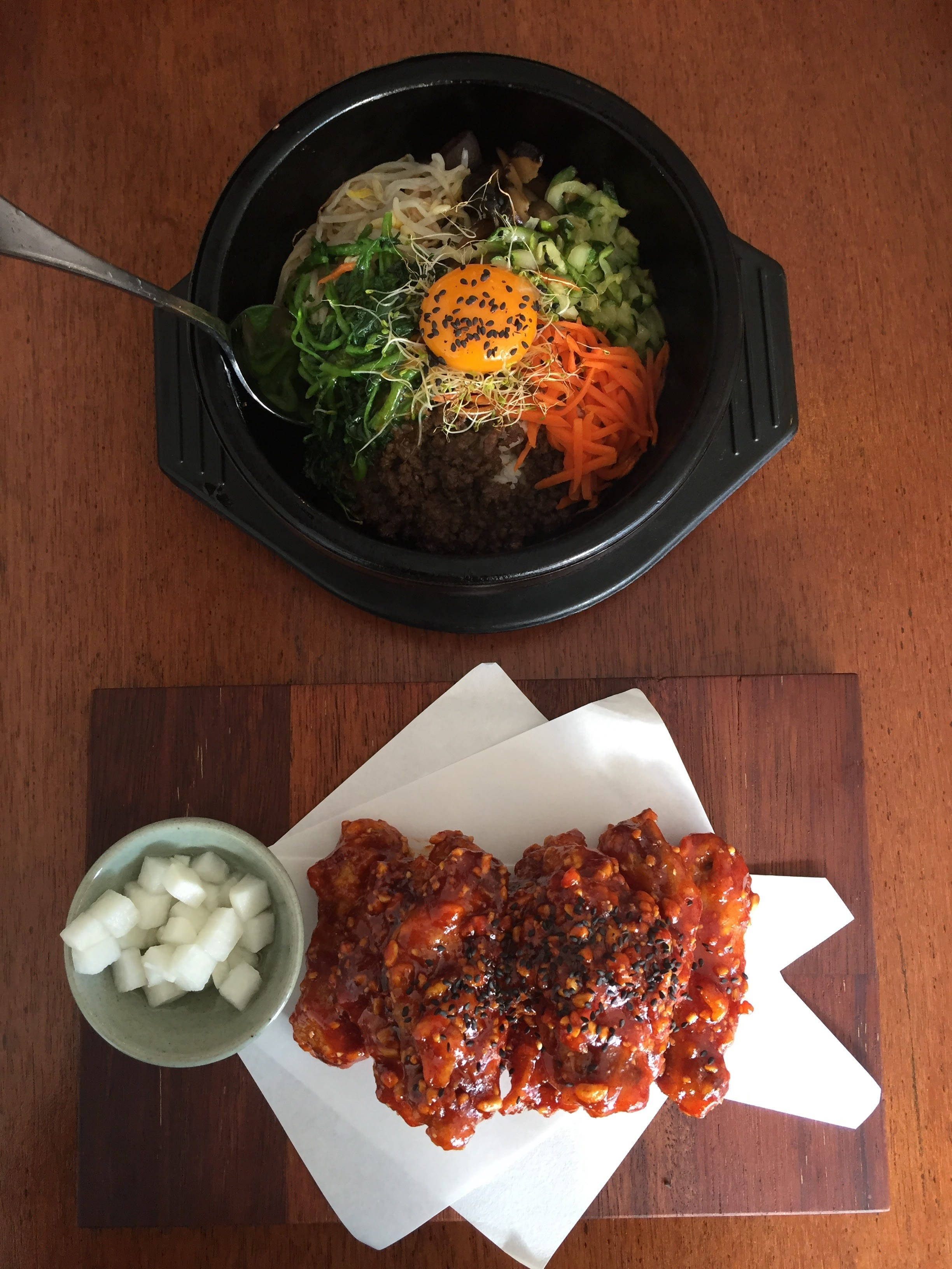 烹飪 桌布 壁紙 — 俯視角度下，韓國石鍋拌飯搭配色彩鮮豔的蔬菜和生蛋黃，旁邊是辣味雞肉和蘿蔔，放在木桌上。