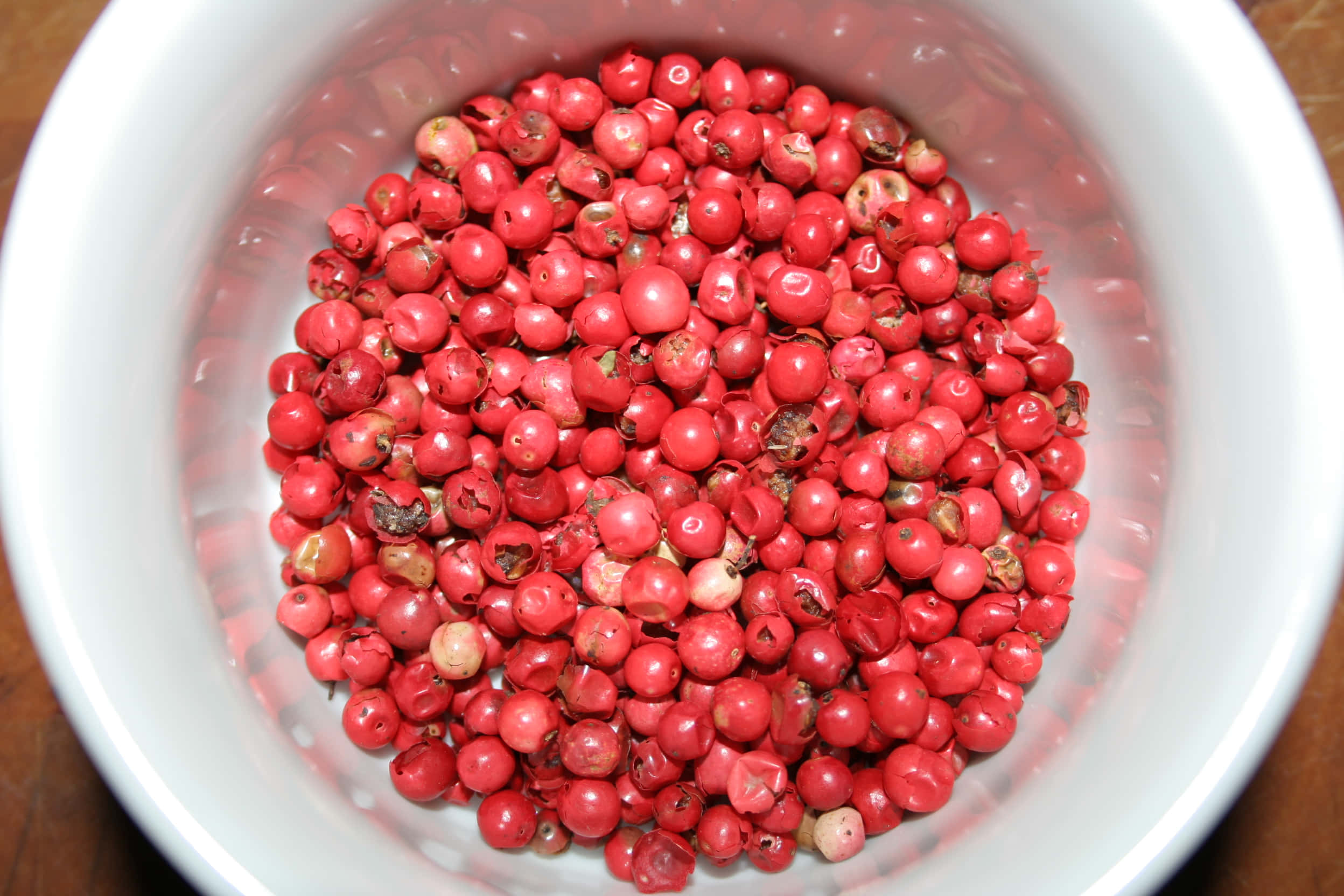 Pink Peppercorn 桌布 壁紙 — 俯視角度拍攝，木質表面上一個白色陶瓷碗裝滿了鮮紅色的胡椒粒。