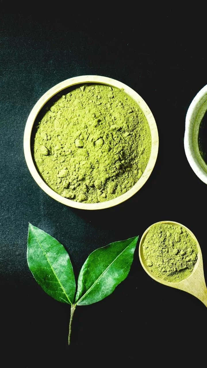 Matcha 桌布 壁紙 — 俯視角度拍攝，深色表面上木碗和木勺中盛有鮮綠色抹茶粉，旁邊放有新鮮茶葉。