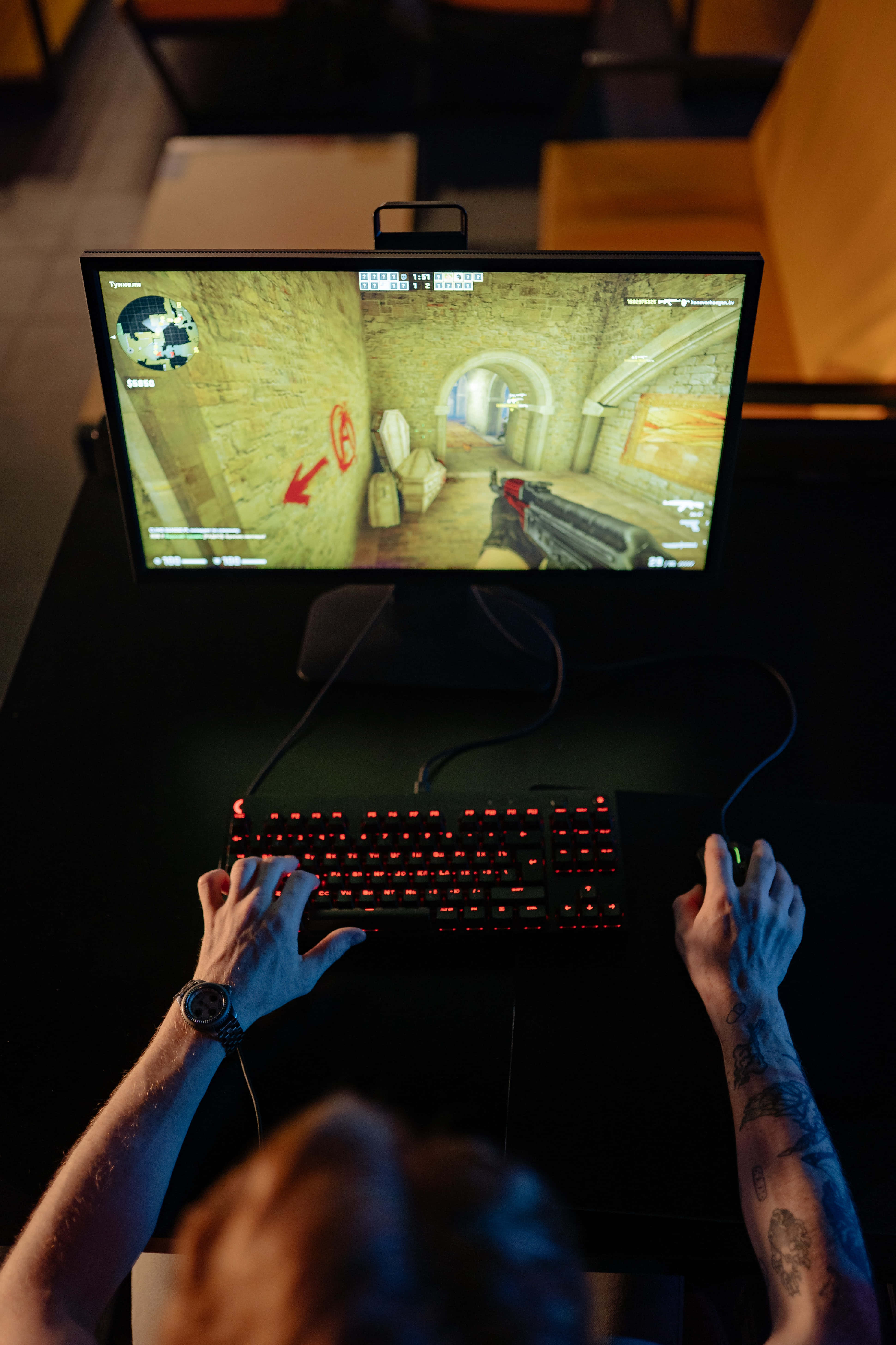 Gaming Pc 桌布 壁紙 — 俯視角度拍攝一位有刺青手臂的玩家，在昏暗房間中於發光紅色鍵盤上激烈地玩第一人稱射擊遊戲。