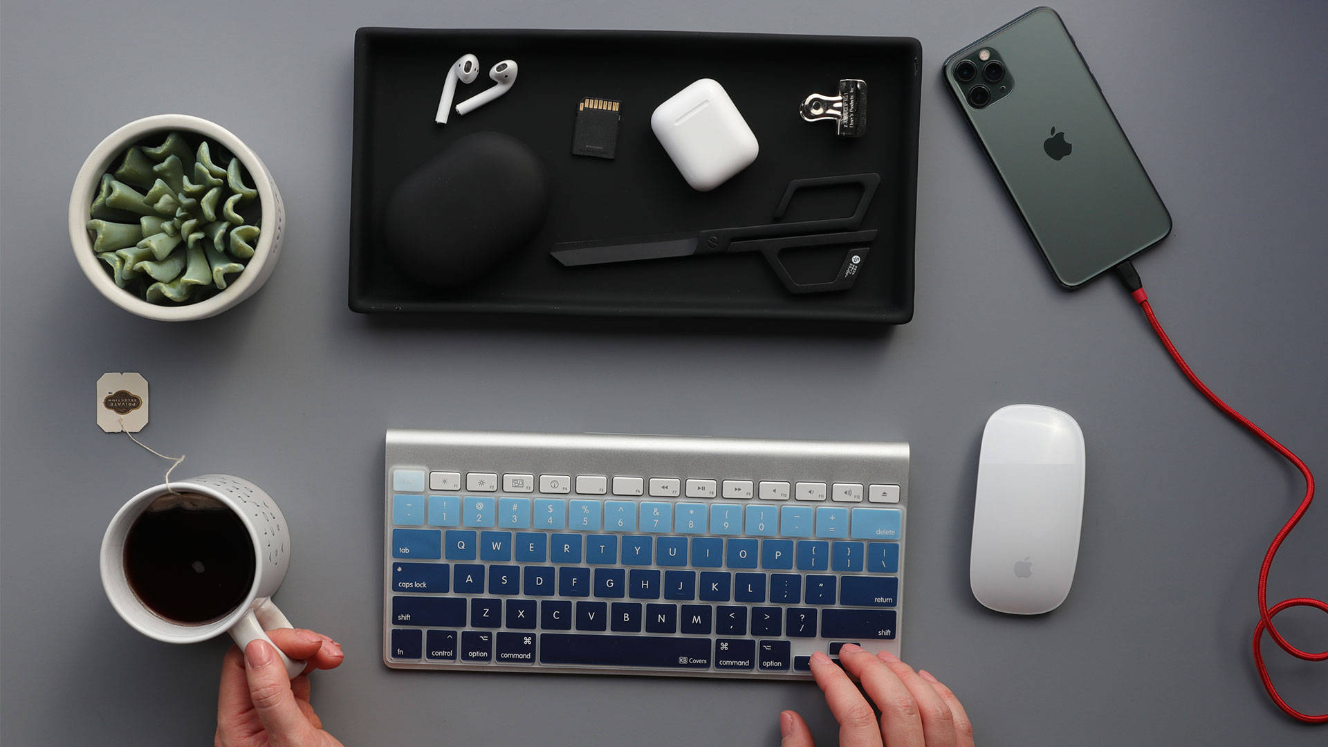 Computer Keyboard 桌布 壁紙 — 俯視角度拍攝雙手在灰色桌上的銀色鍵盤打字，周圍有科技產品和一杯茶。
