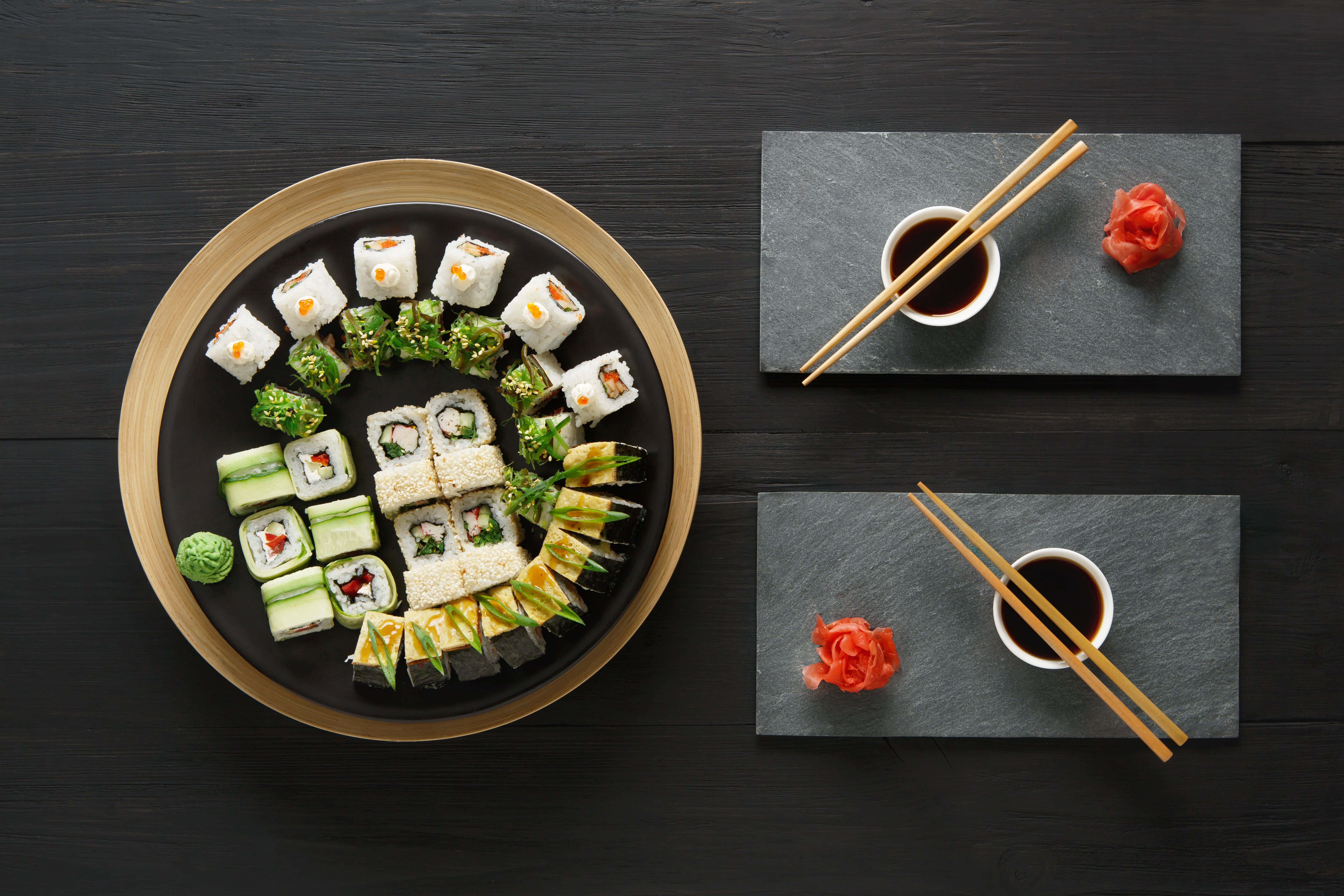 Food Photography 桌布 壁紙 — 俯視角度拍攝黑色盤子上放滿各式壽司卷和生魚片，以芥末和生薑點綴，置於深色木桌上。