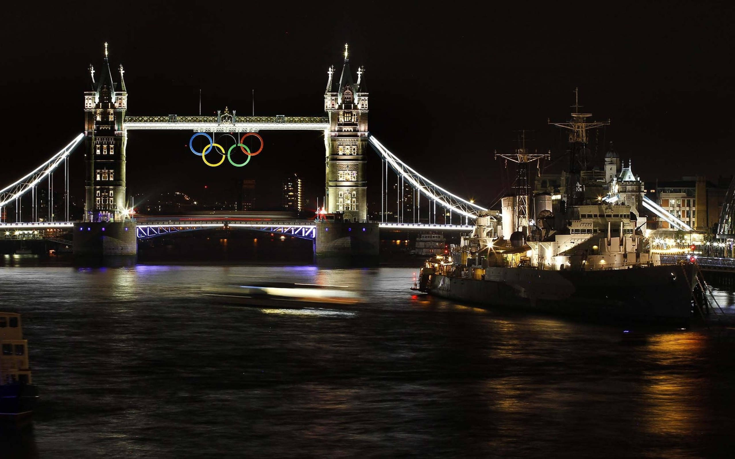 England 桌布 壁紙 — 倫敦塔橋在夜晚亮燈，奧運五環在上方閃耀，倒映在泰晤士河上，旁邊有一艘歷史戰艦。
