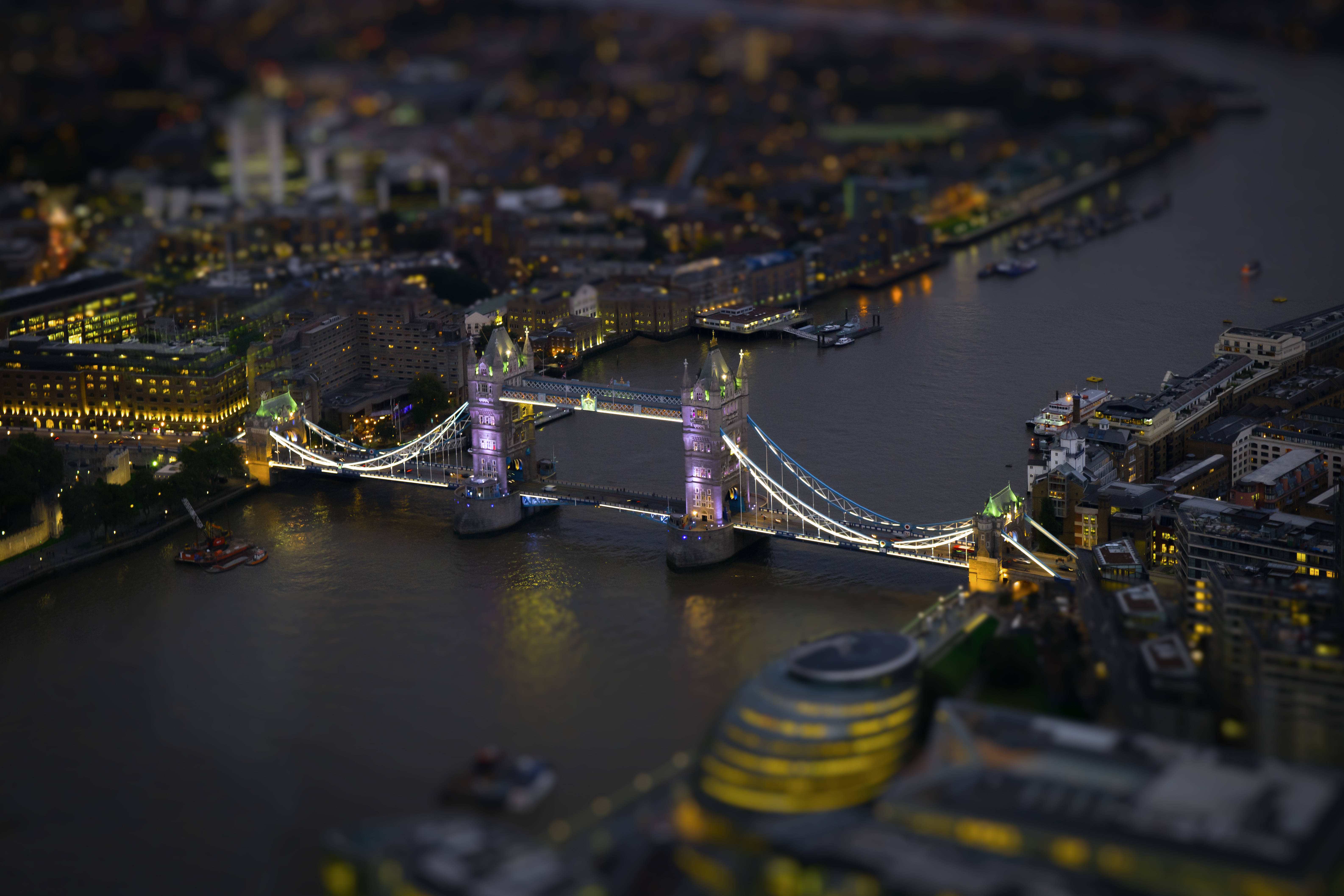 United Kingdom 桌布 壁紙 — 倫敦塔橋在黃昏時的微縮移軸視角，發光的燈光倒映在河面上。