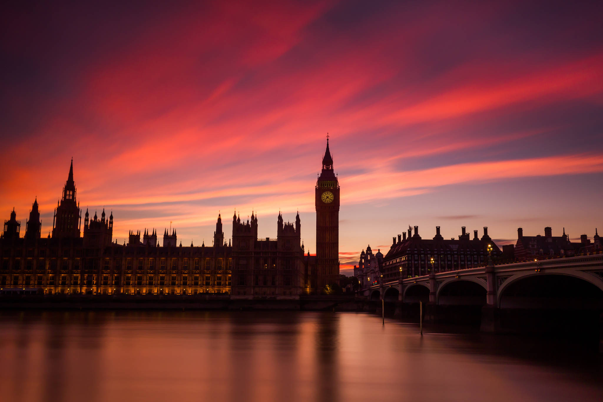 England 桌布 壁紙 — 倫敦標誌性的大笨鐘和國會大廈在火紅橙色的日落天空襯托下形成剪影，倒映在平靜的泰晤士河中。