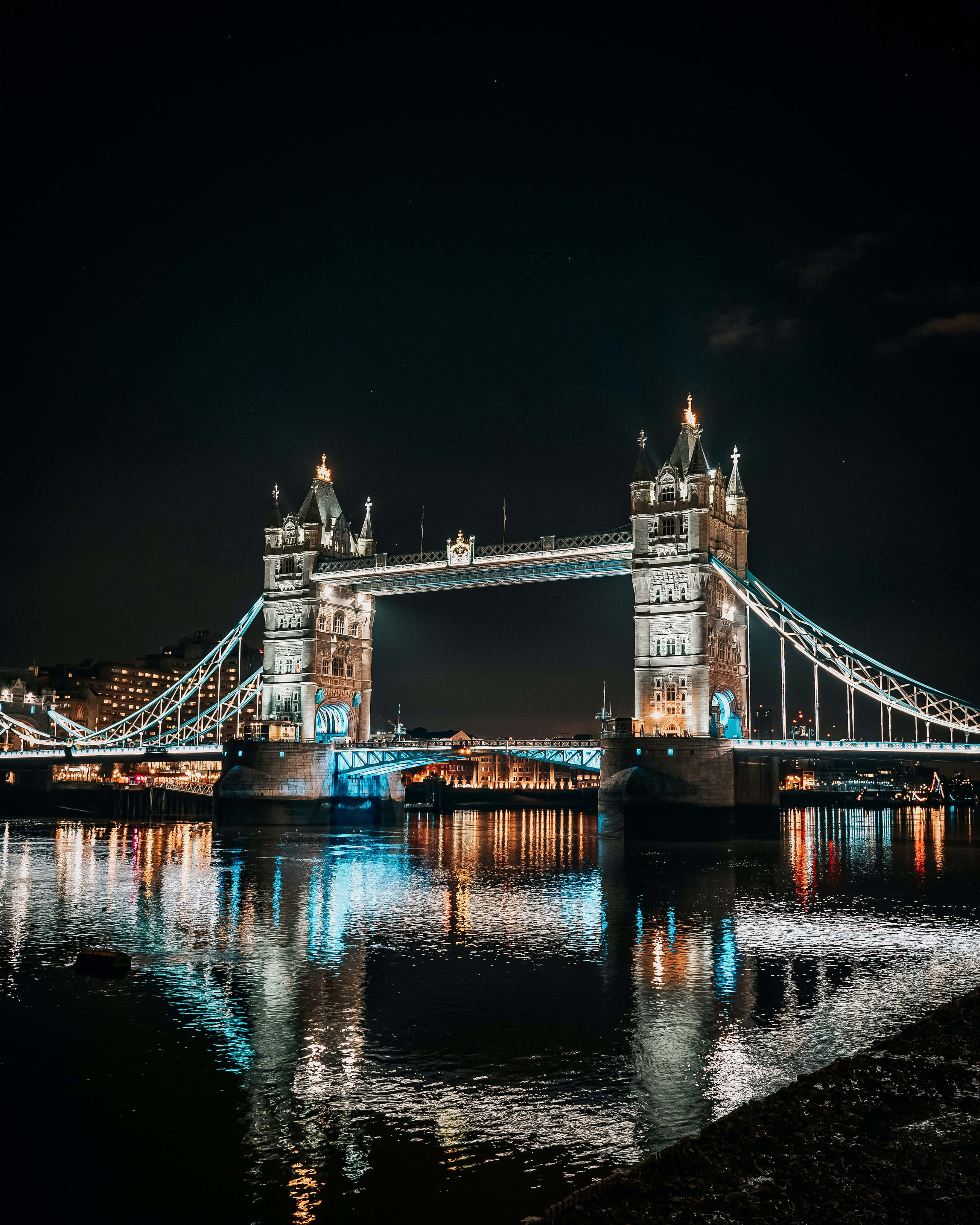 London iPhone 桌布 壁紙 — 倫敦的塔橋在夜間閃爍著藍色和白色的燈光，倒映在泰晤士河上。