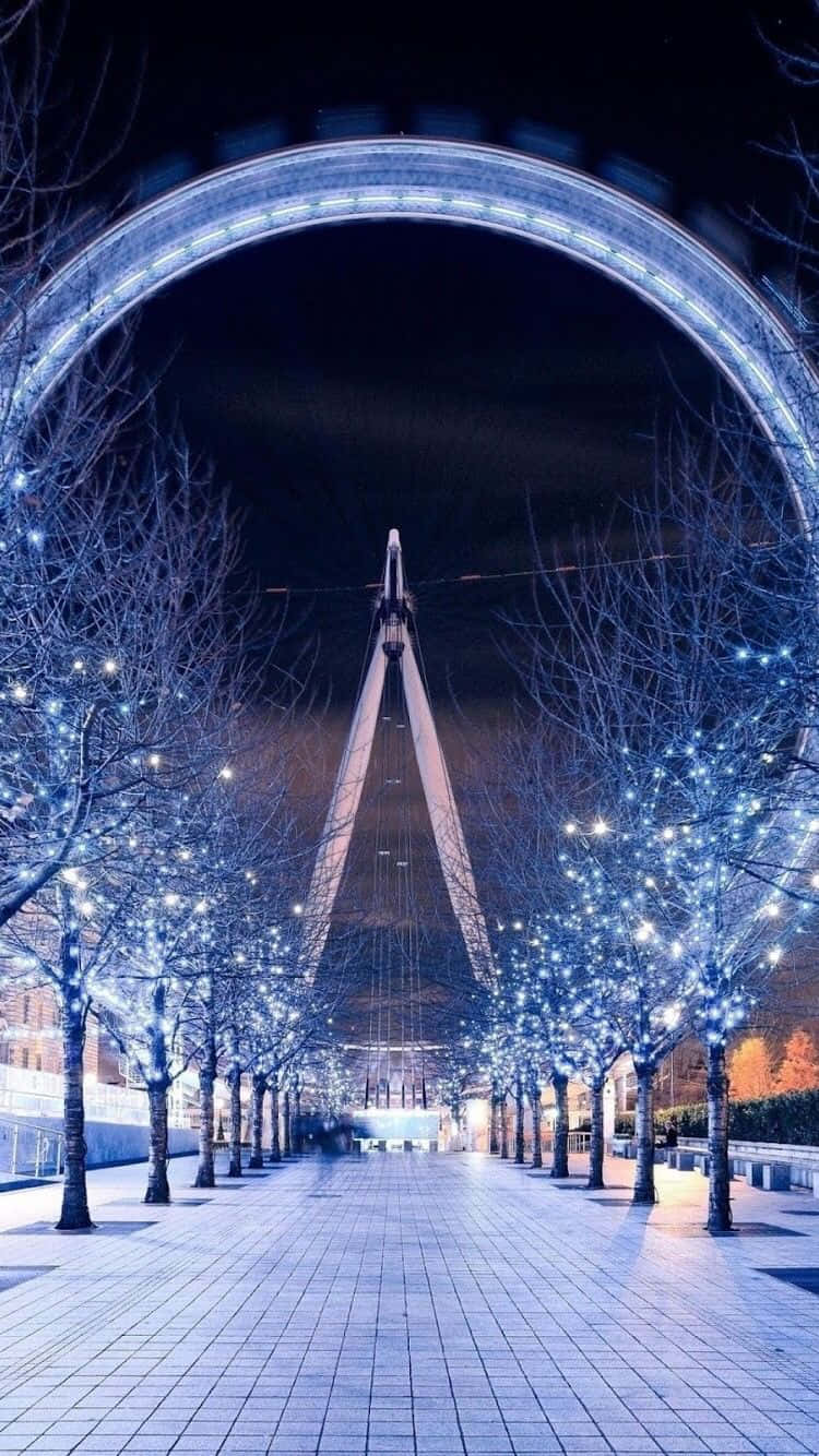 England 桌布 壁紙 — 倫敦眼在夜晚的耀眼景色，由對稱的裸樹大道框住，樹上裝飾著閃爍的藍色燈光。