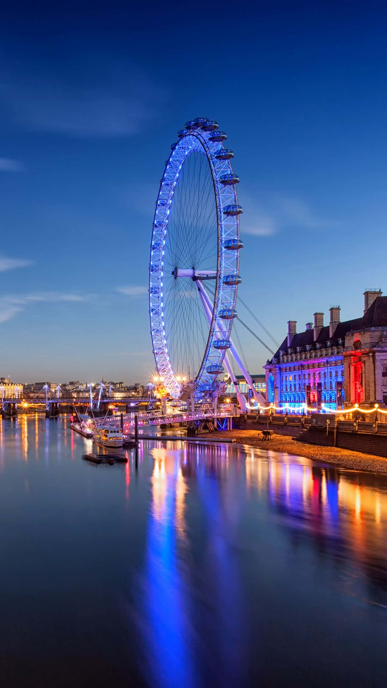 London iPhone 桌布 壁紙 — 倫敦眼在暮色天空下散發出鮮豔的藍色光芒，其倒影在平靜的泰晤士河上閃爍。