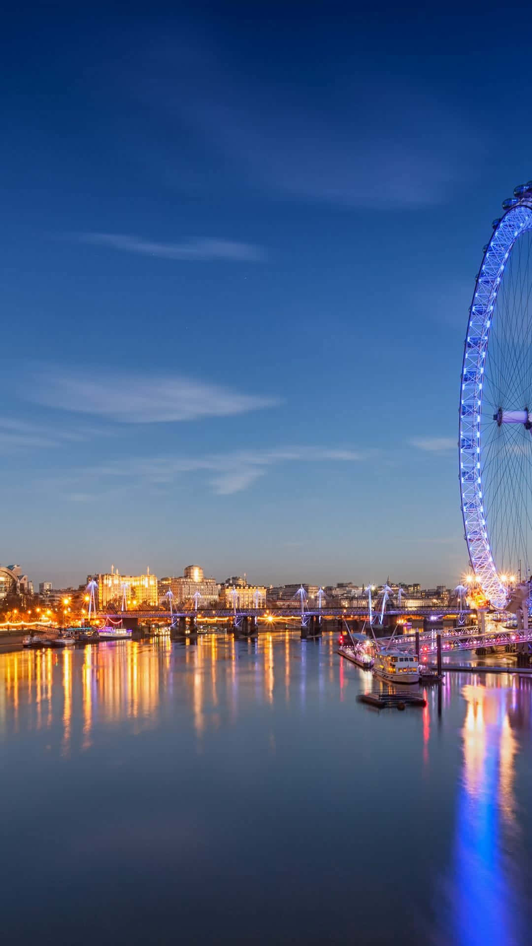 London iPhone 桌布 壁紙 — 倫敦眼在暮色天空中發出藍色光芒，城市燈光倒映在下方平靜的泰晤士河上。