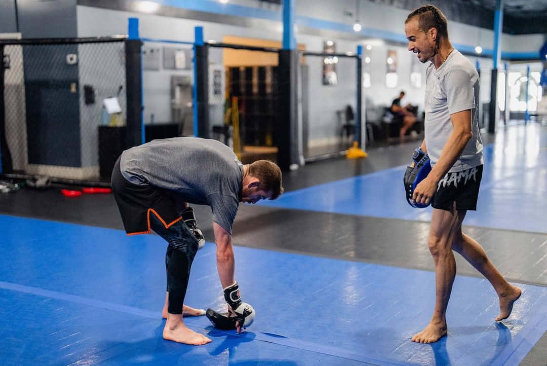 MMA 桌布 壁紙 — 健身房裡有兩個男人，一個彎腰調整手套，另一個微笑著走過藍色墊子。