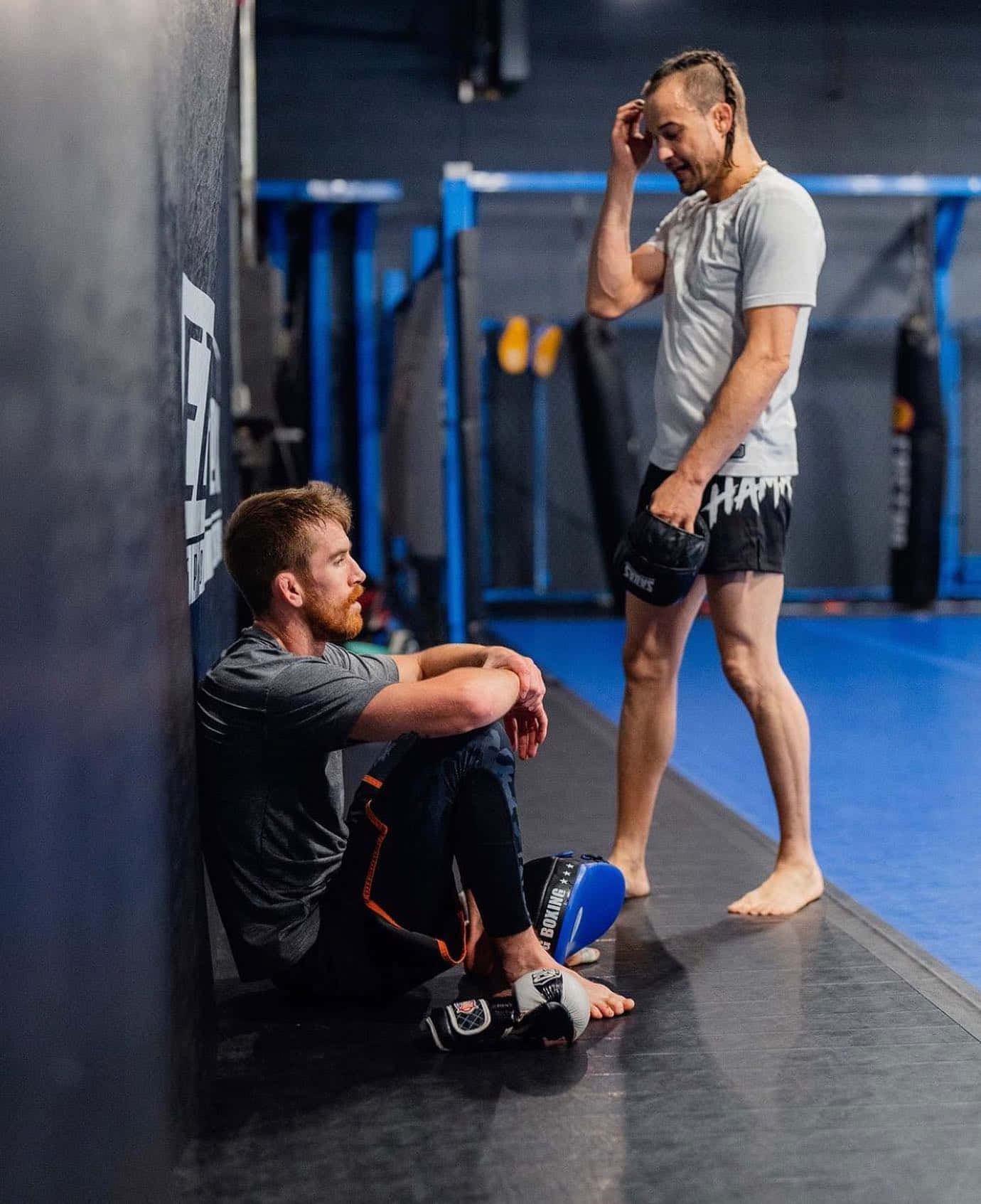 UFC 桌布 壁紙 — 健身房裡有兩名拳手，一人筋疲力盡地靠著軟墊牆坐著，另一人站在附近拿著手套。
