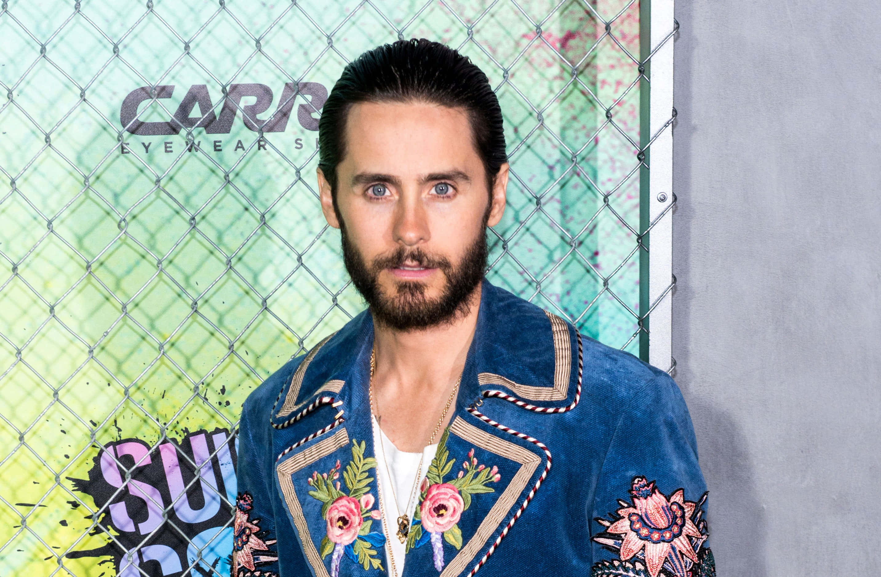 jared leto 桌布 壁紙 — 傑克·葛倫霍自信地站在一個塗鴉噴灑的鐵絲網圍欄前，身穿鮮豔的藍色花卉刺繡外套。