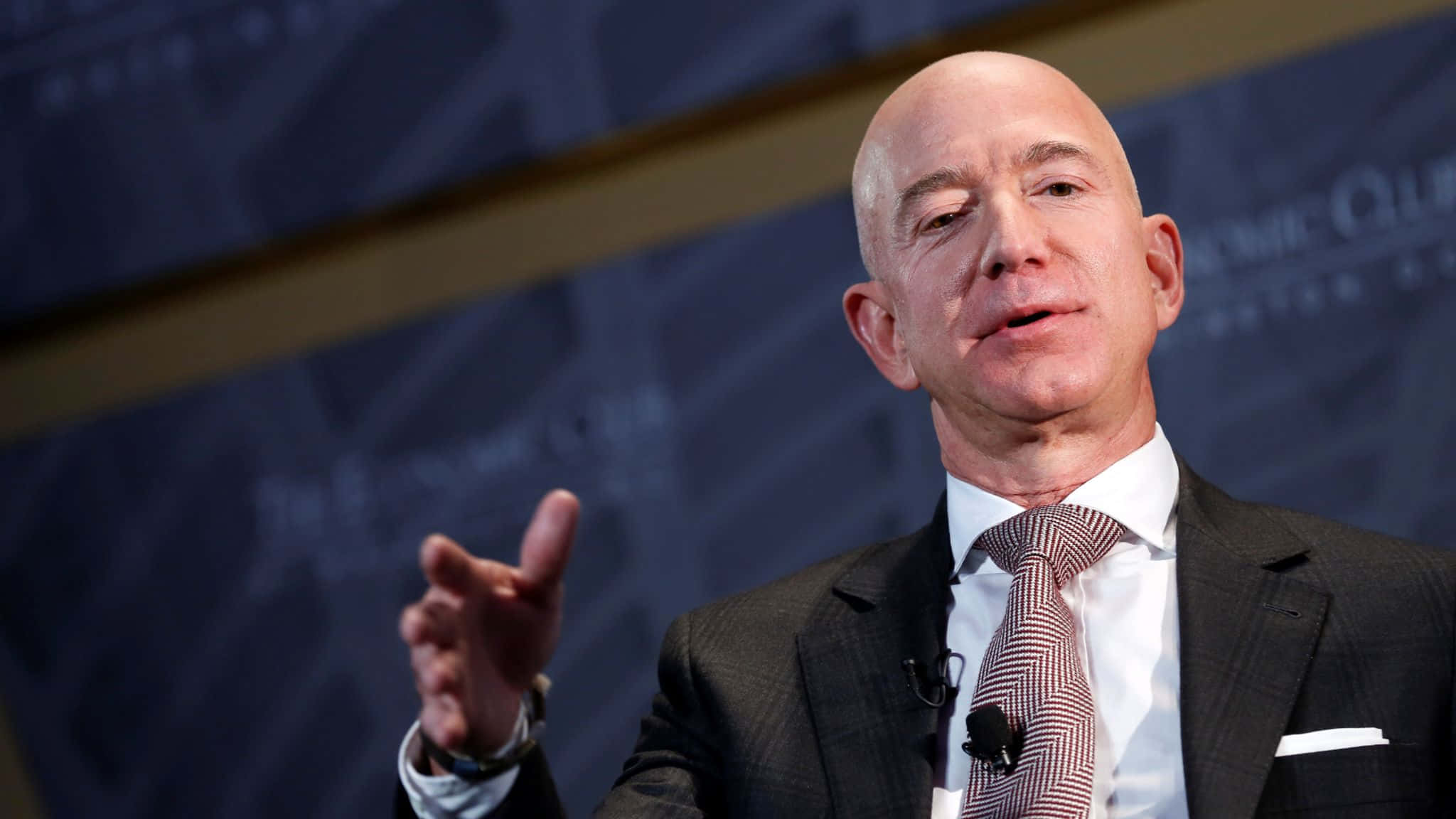 Jeff Bezos 桌布 壁紙 — 傑夫·貝佐斯在正式活動中發言，身穿深色西裝和圖案領帶，背景為模糊的藍色。