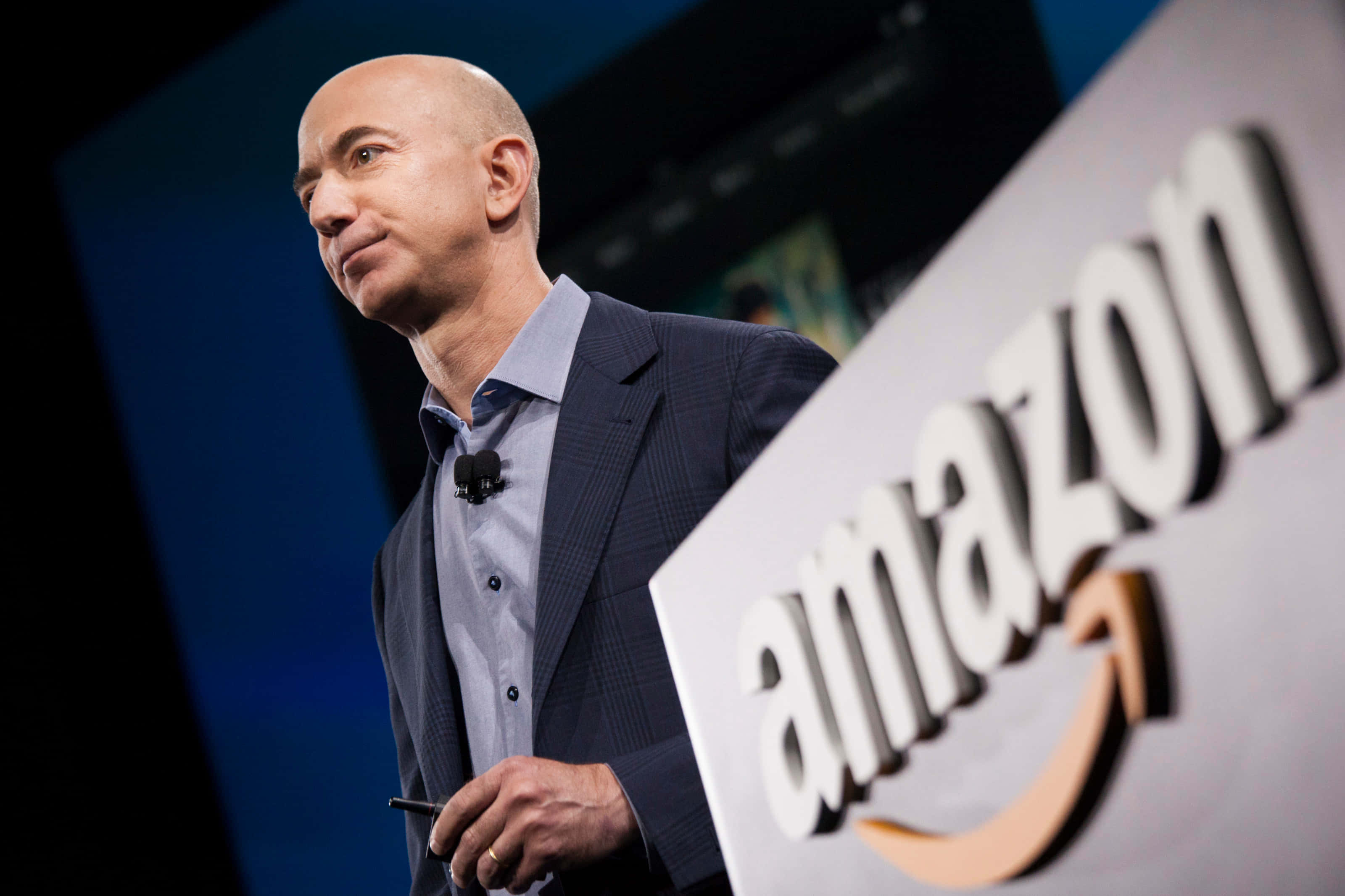 Jeff Bezos 桌布 壁紙 — 傑夫·貝佐斯在舞台燈光下的企業活動中自信地站在一個大型亞馬遜標誌旁邊。