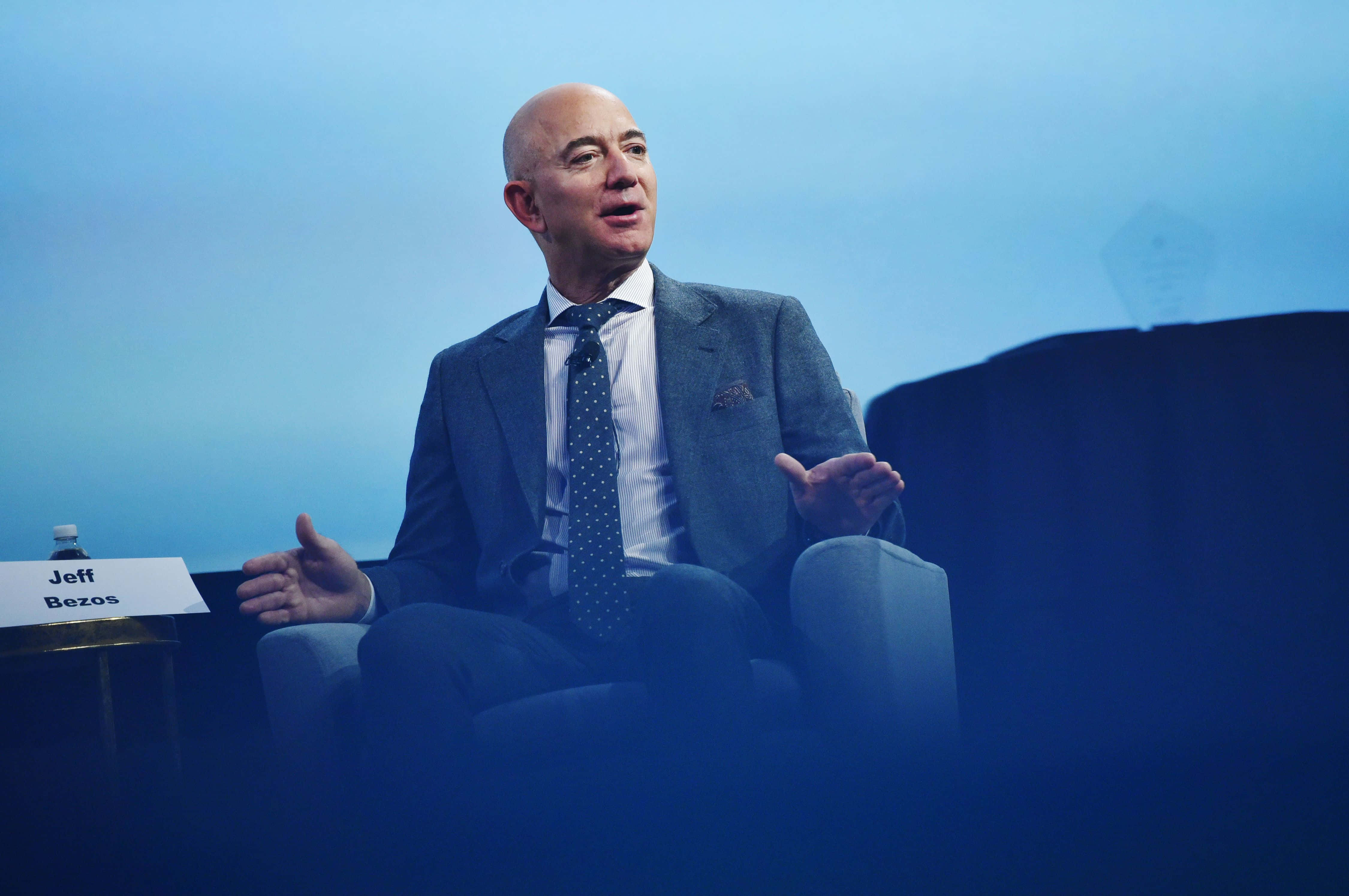 Jeff Bezos 桌布 壁紙 — 傑夫·貝佐斯在藍色燈光的會議室中，穿著灰色西裝和波點領帶，生動地說話。