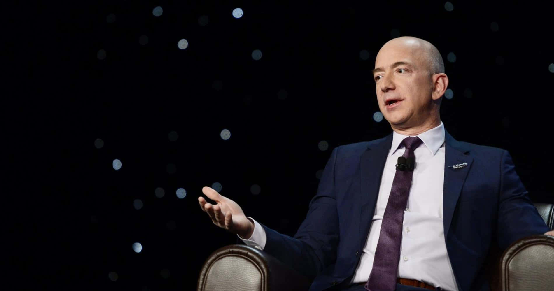 Jeff Bezos 桌布 壁紙 — 傑夫·貝佐斯坐在椅子上，背景昏暗帶有散景燈光，穿著正式服裝說話時做手勢。