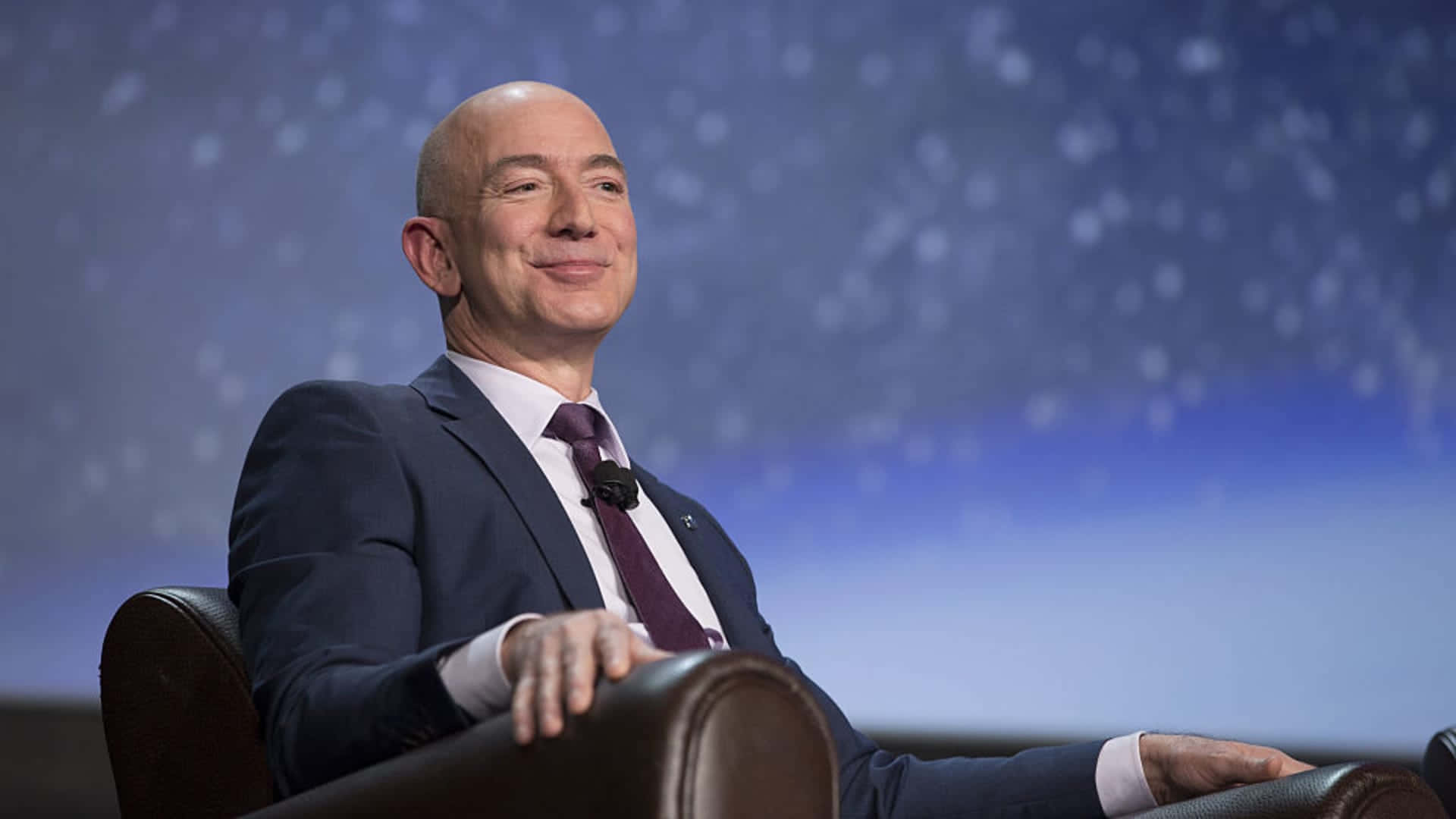 Jeff Bezos 桌布 壁紙 — 傑夫·貝佐斯放鬆地坐在皮革椅上，背景是星光閃爍的藍色，面帶溫柔微笑。