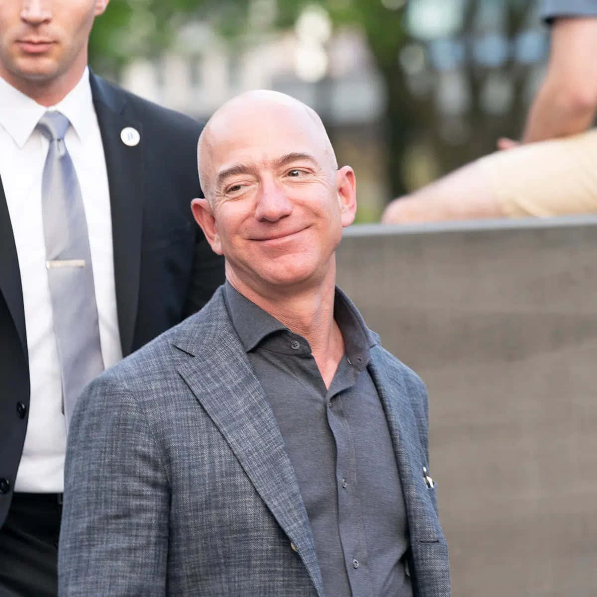 Jeff Bezos 桌布 壁紙 — 傑夫·貝佐斯穿著灰色西裝溫暖微笑，身後站著一位身穿深色西裝的保安人員在戶外。