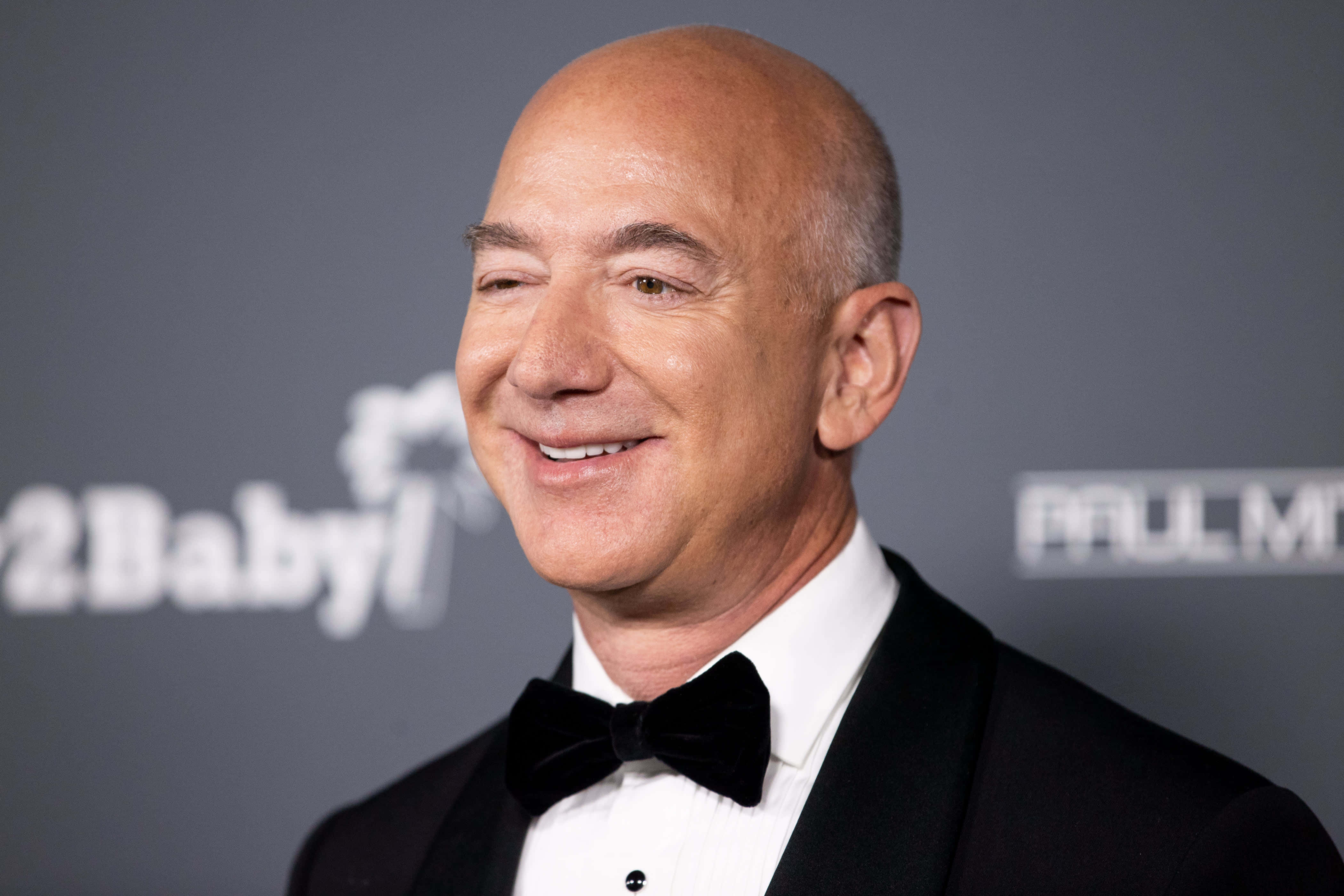 Jeff Bezos 桌布 壁紙 — 傑夫·貝佐斯穿著黑色燕尾服和領結，在帶有標誌的灰色活動背景前溫暖地微笑。