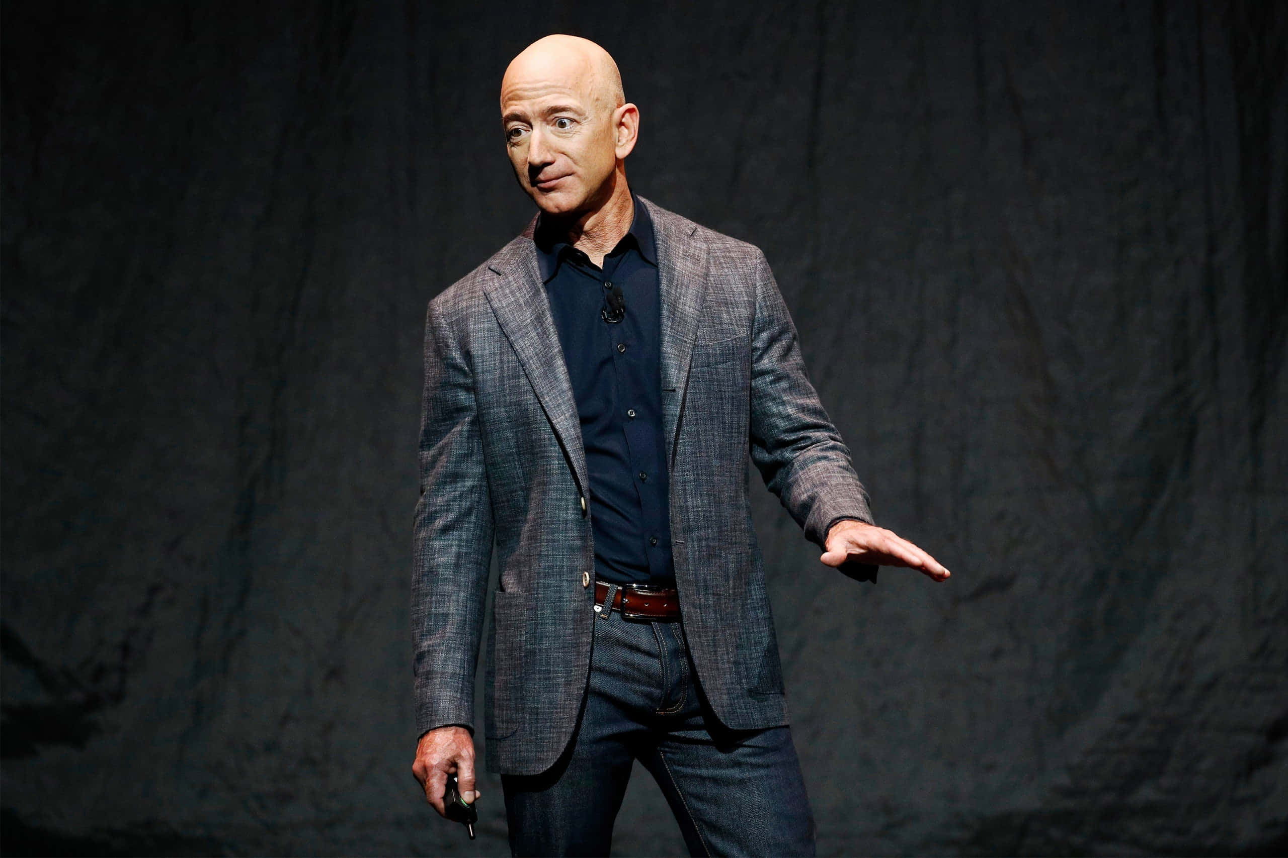 Jeff Bezos 桌布 壁紙 — 傑夫·貝佐斯自信地站在舞台上，身穿格紋西裝外套，背景昏暗，在明亮的聚光燈下演講時做手勢。