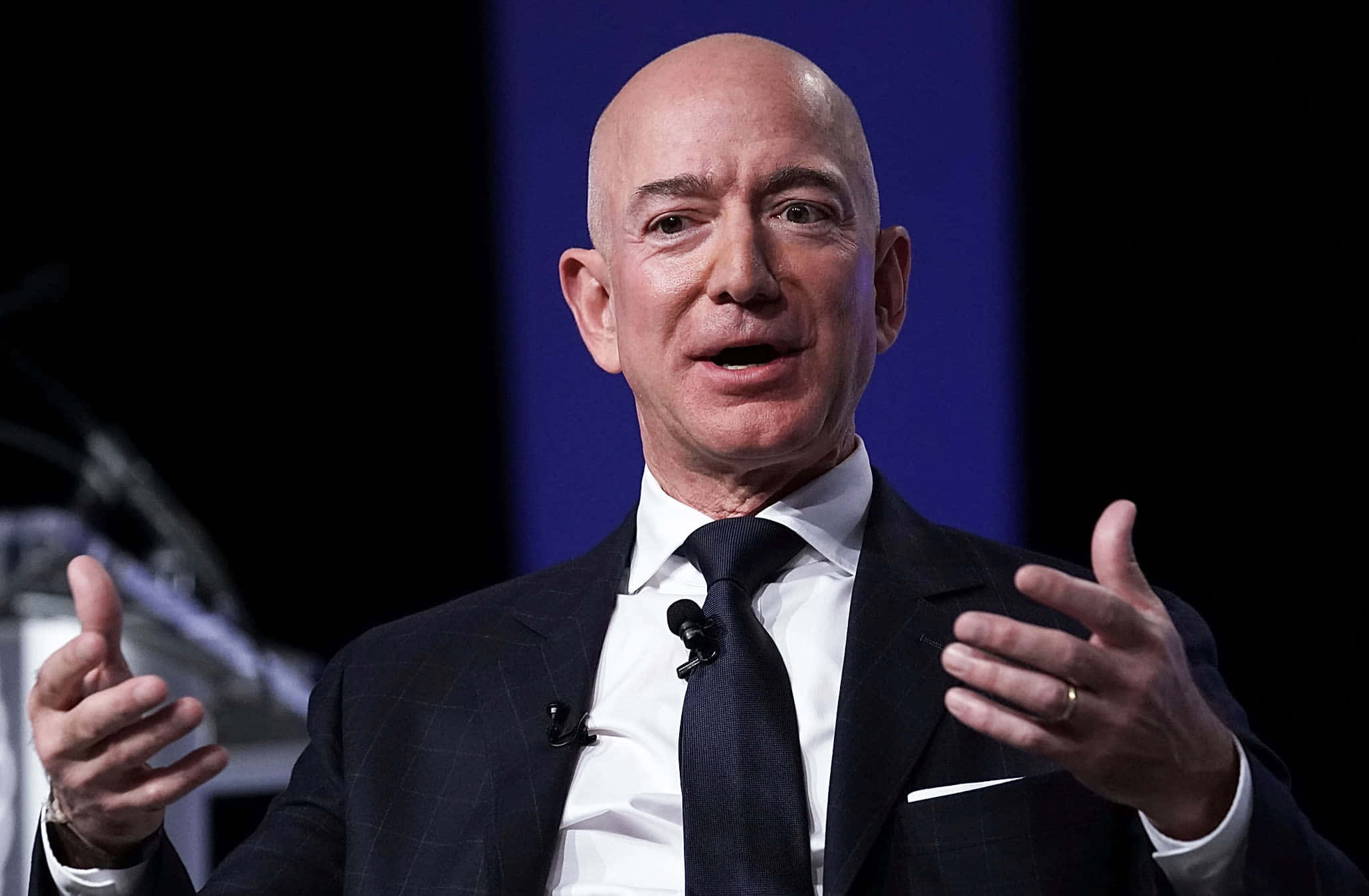 Jeff Bezos 桌布 壁紙 — 傑夫·貝佐斯身穿深色西裝，在藍色背景前，雙手在句子中間做手勢，充滿激情地演講。