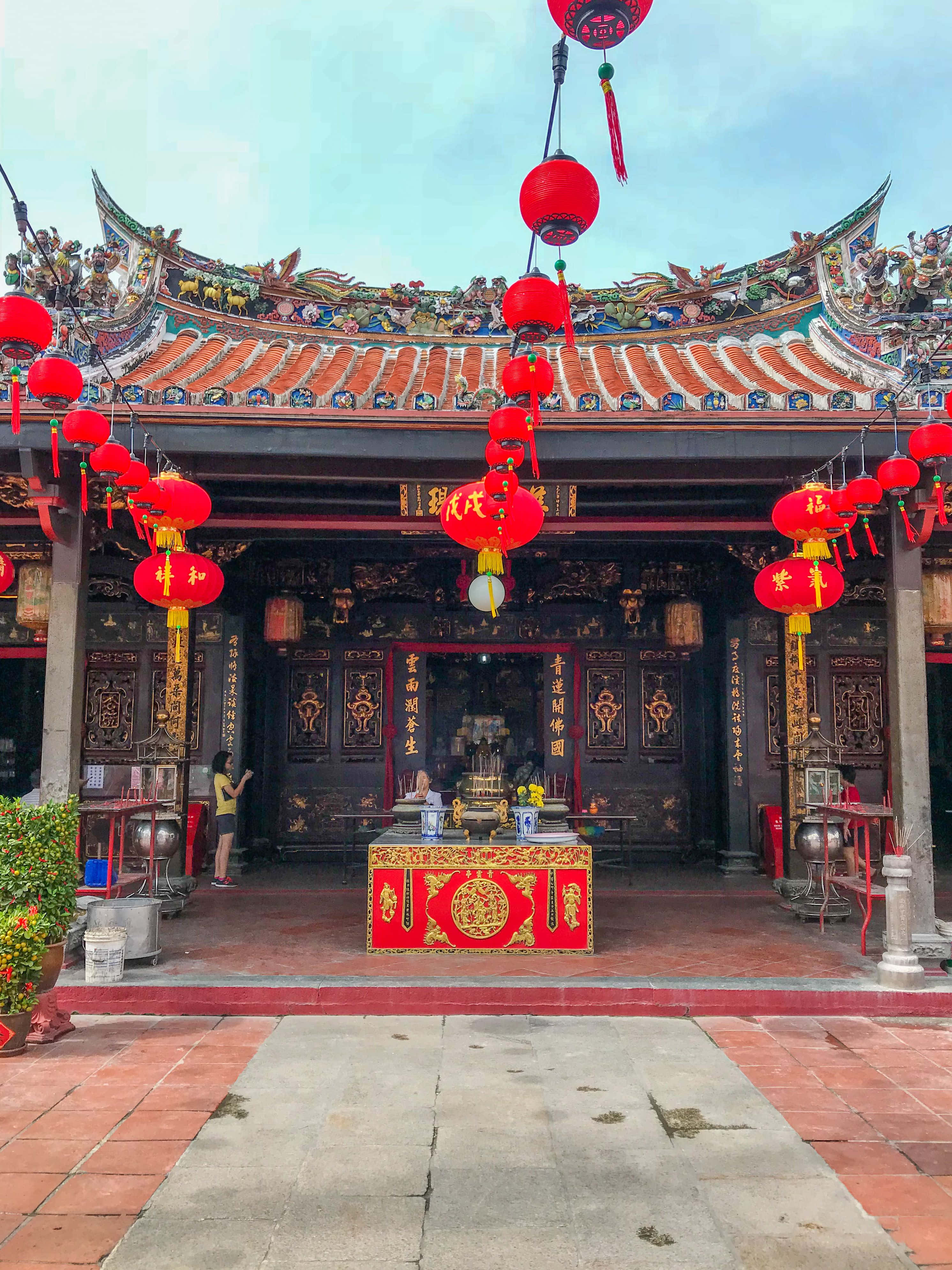 春節 桌布 壁紙 — 傳統中國寺廟，擁有華麗的瓦片屋頂和紅色燈籠，懸掛在紅色和金色的祭壇上方，天空晴朗。