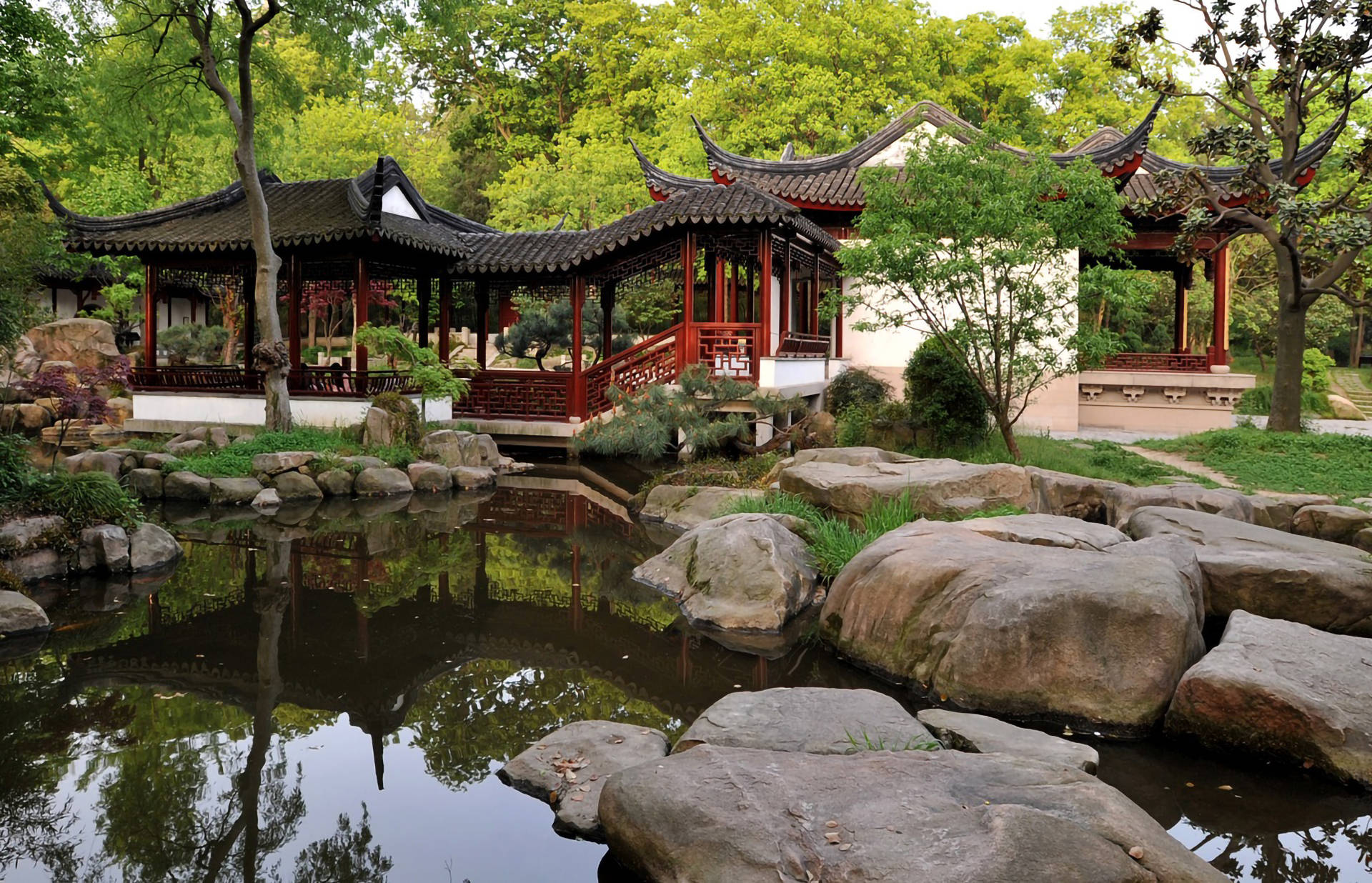 Suzhou 桌布 壁紙 — 傳統中式亭台樓閣，曲線屋頂倒映在平靜的池塘中，四周環繞著茂密的綠樹和長滿青苔的岩石。
