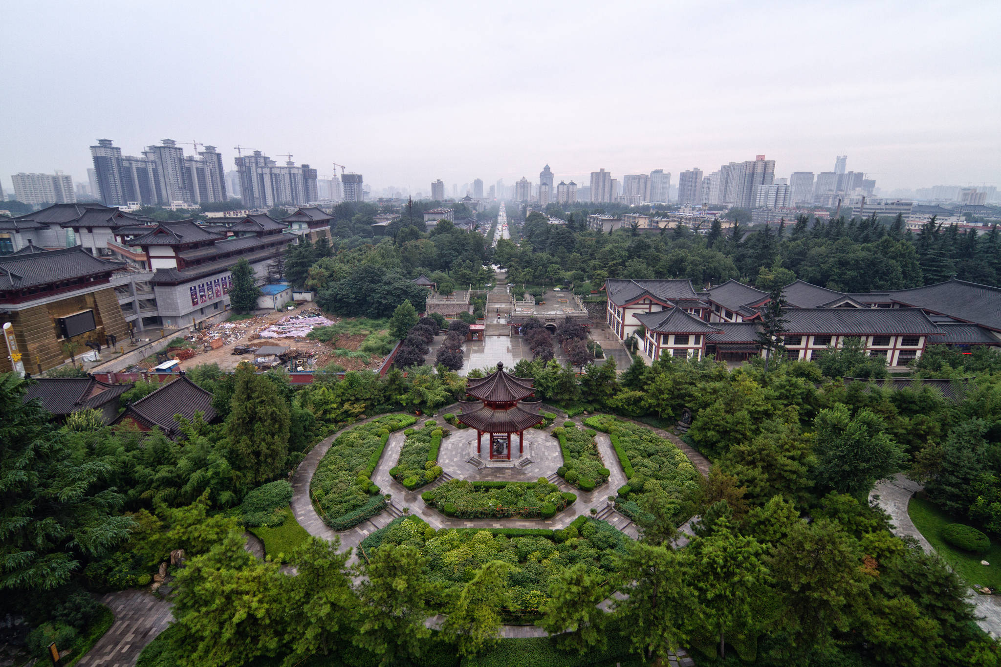 Xian 桌布 壁紙 — 傳統中式亭台樓閣和花園的鳥瞰圖，周圍環繞著茂密的綠植，遠處朦朧中可見現代城市天際線。