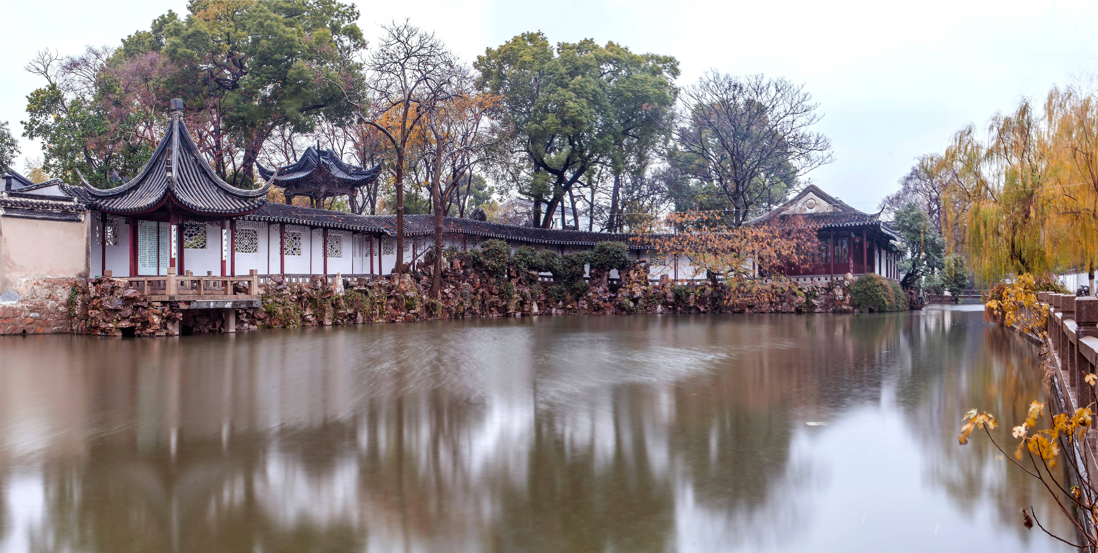 Suzhou 桌布 壁紙 — 傳統中式亭閣的彎曲屋頂倒映在平靜的水面上，周圍環繞著秋樹。