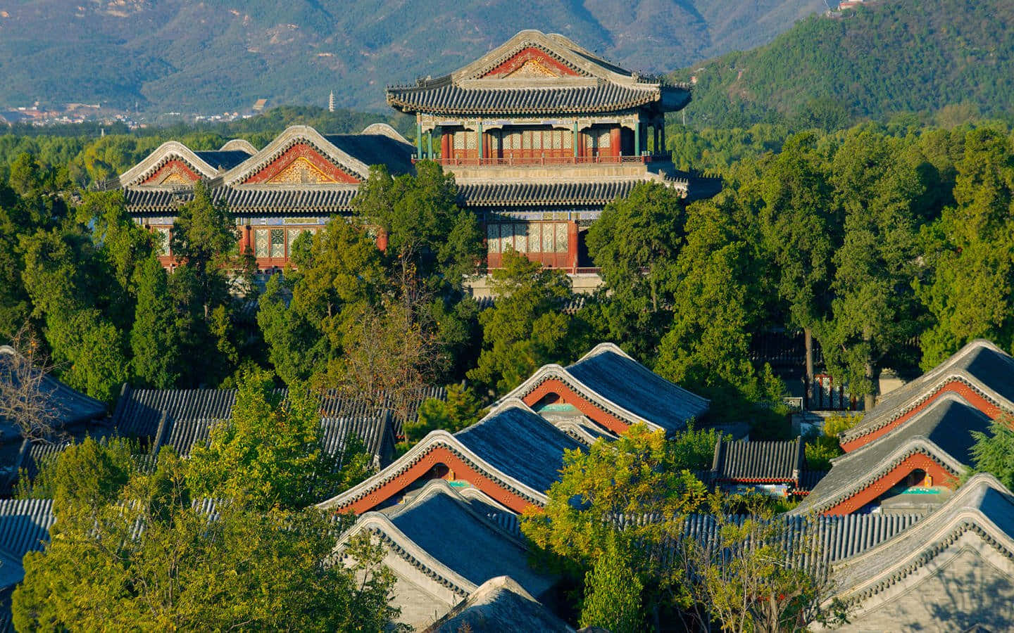 樹木 桌布 壁紙 — 傳統中式宮殿建築，擁有彎曲的瓦片屋頂，坐落在茂密的綠樹之間，以山巒為背景。