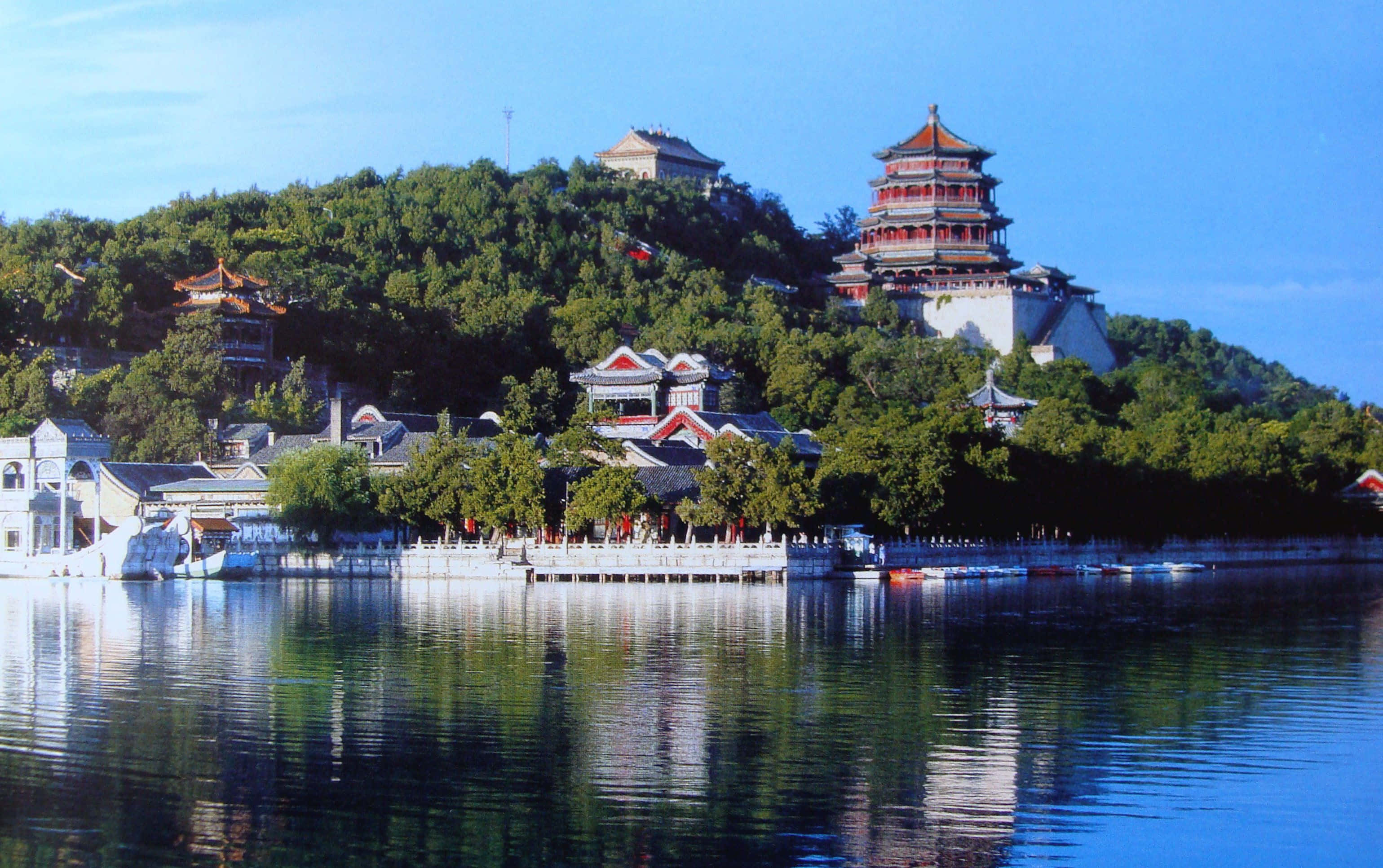 山景 桌布 壁紙 — 傳統中式寶塔和亭台樓閣坐落在鬱鬱蔥蔥的青山上，在晴朗的天空下完美地倒映在平靜的藍色水面上。