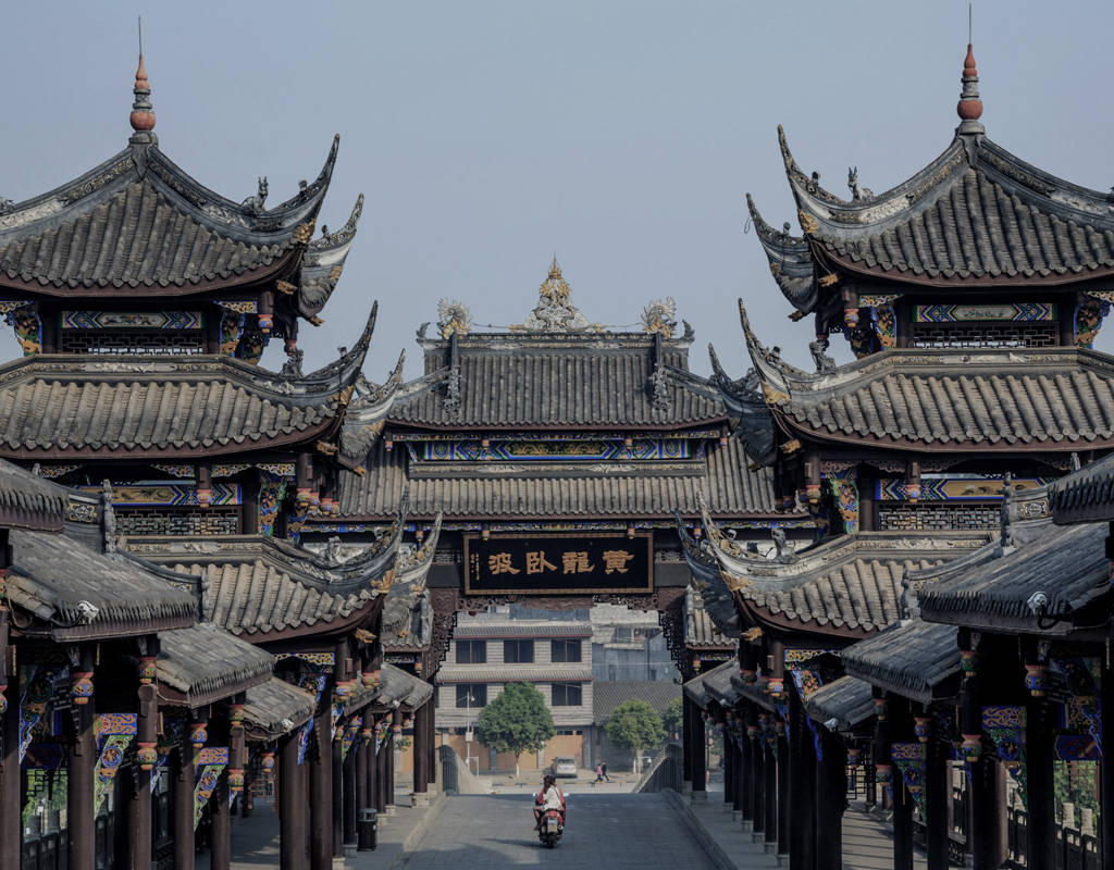 Chengdu 桌布 壁紙 — 傳統中式建築的對稱景觀，擁有華麗的瓦片屋頂，以及一名孤獨的摩托車騎士在石橋上行駛，天空朦朧。