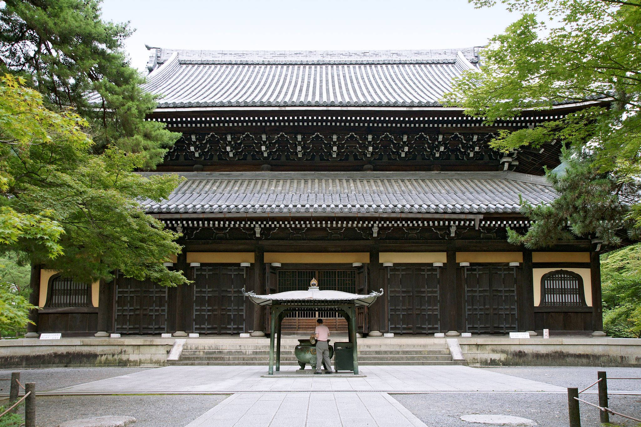 京都 桌布 壁紙 — 傳統日本寺廟，深色木結構和瓦片屋頂，周圍環繞著茂密的綠樹，天空多雲。