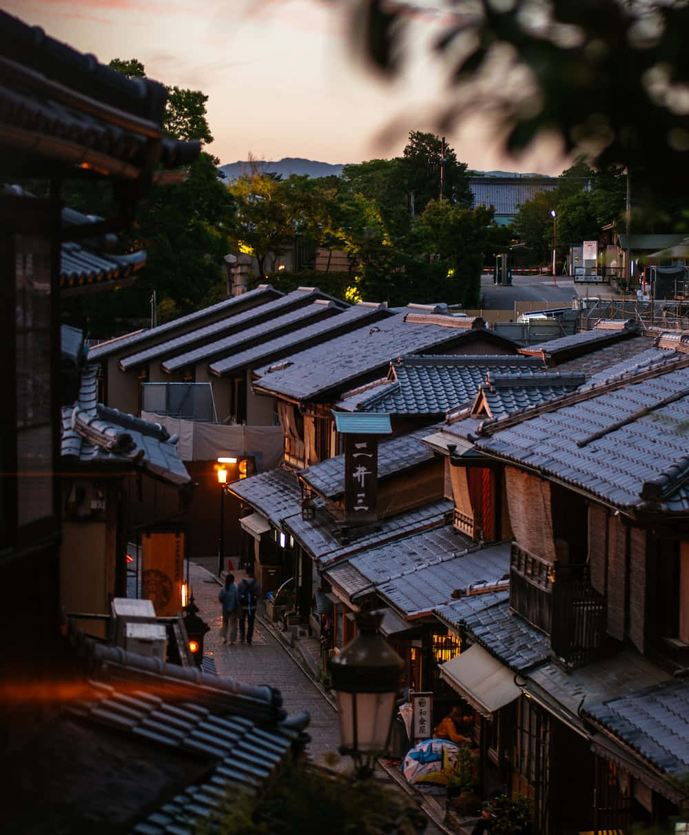 日本 桌布 壁紙 — 傳統日本屋頂和燈籠照亮的小巷，遠山環繞的寧靜黃昏景色。