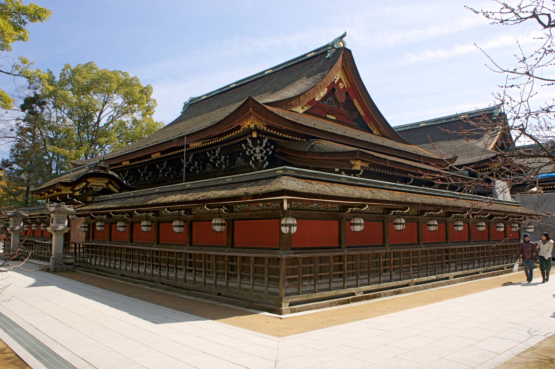 傳統日本神社，紅色牆壁和彎曲屋頂，在藍天下，有人在附近行走。