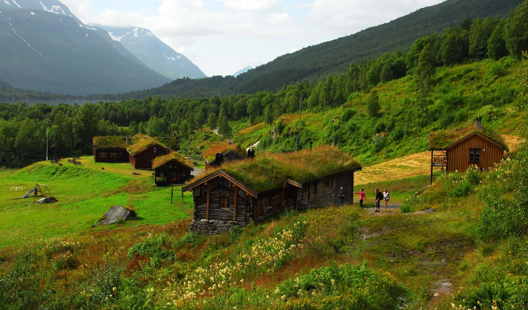 Old Farm 桌布 壁紙 — 傳統木屋，屋頂覆蓋著草，坐落在鬱鬱蔥蔥的綠色山谷中，周圍環繞著森林覆蓋的山脈，天空部分多雲。
