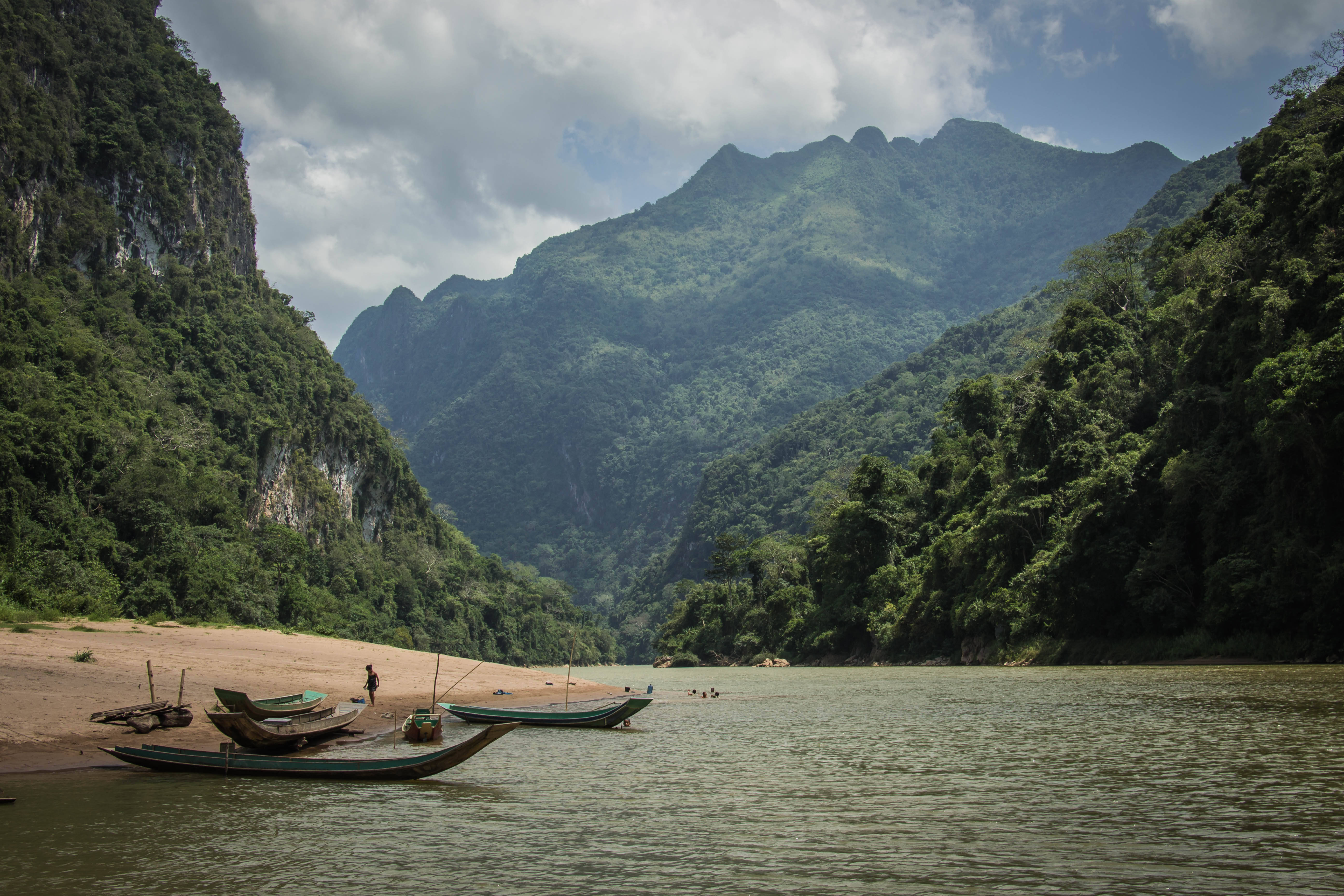 Laos 桌布 壁紙 — 傳統木船停泊在沙質河岸上，背景是雄偉的翠綠山脈，天空部分多雲。