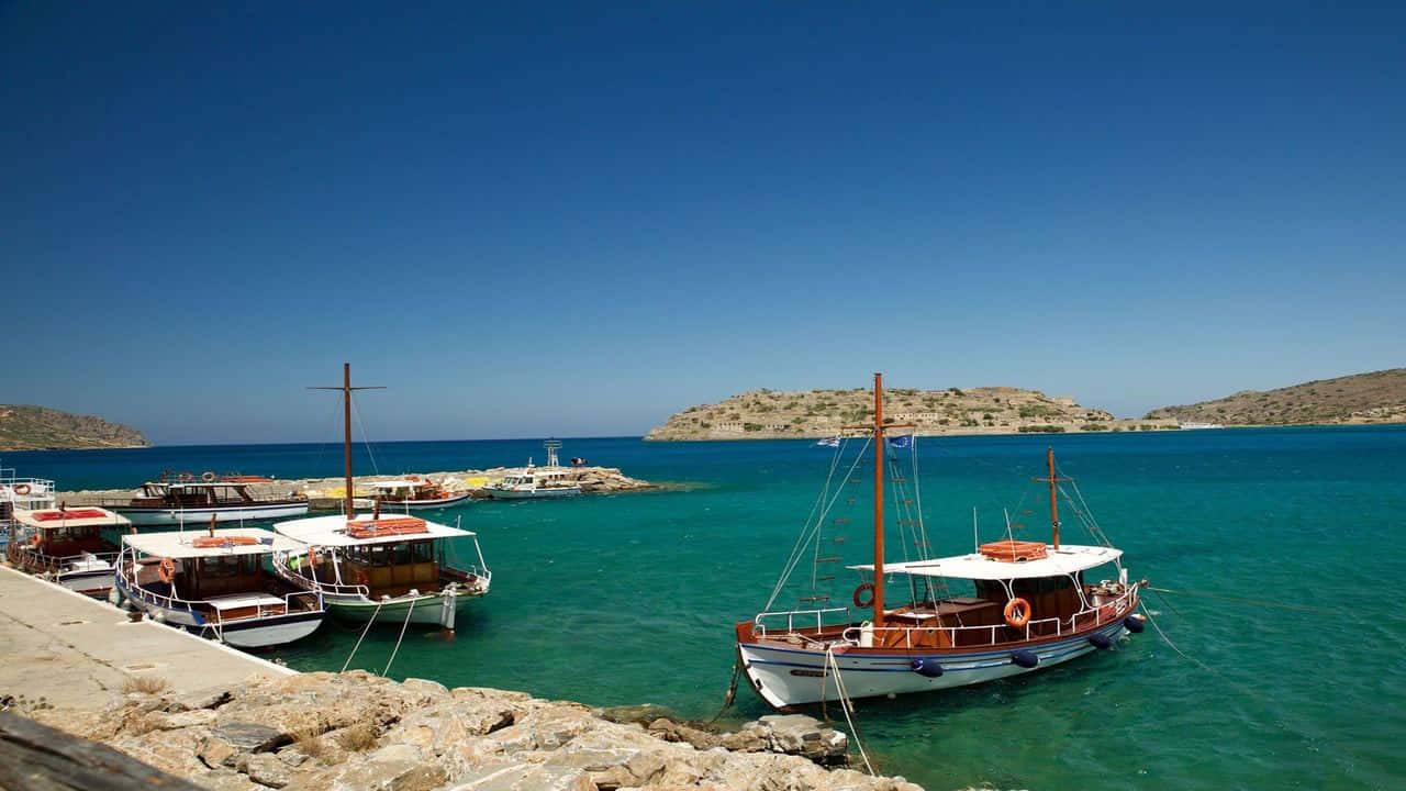 Greek Island 桌布 壁紙 — 傳統木船配白色頂篷在清澈藍天下停泊在岩石碼頭旁的碧綠海水中。
