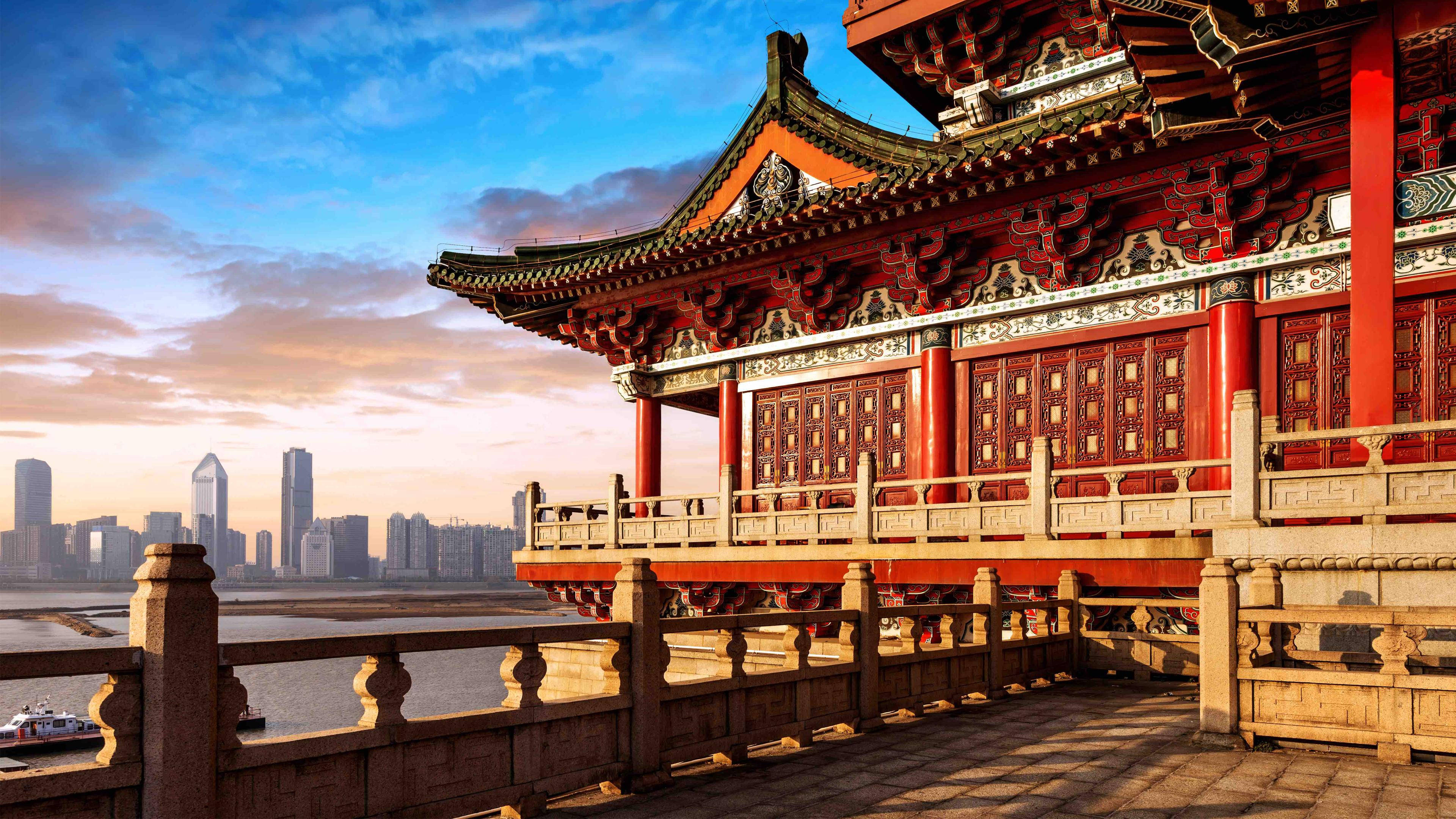 Beijing 桌布 壁紙 — 傳統紅色中式亭閣，曲線屋頂俯瞰河流，映襯現代城市天際線，夕陽西下。