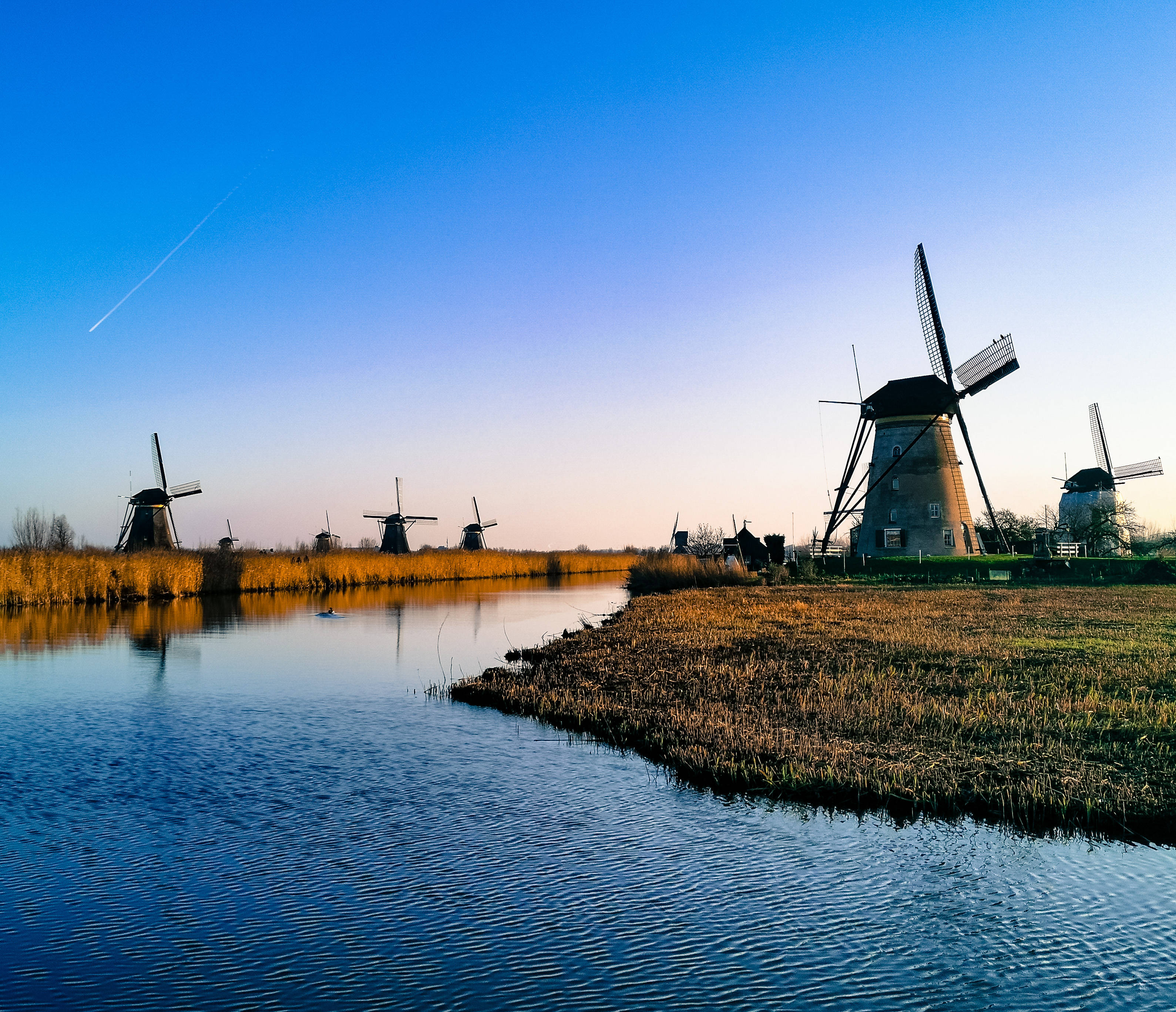 Netherlands 桌布 壁紙 — 傳統荷蘭風車沿著寧靜的運河在日落時分矗立，金色蘆葦和清澈的藍天。