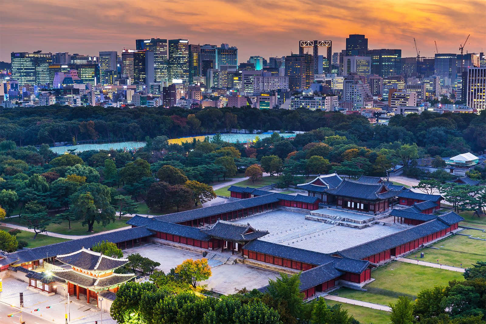 南韓 桌布 壁紙 — 傳統韓國宮殿，紅牆黑瓦，坐落在綠色公園中，以夕陽下閃耀的現代城市天際線為背景。