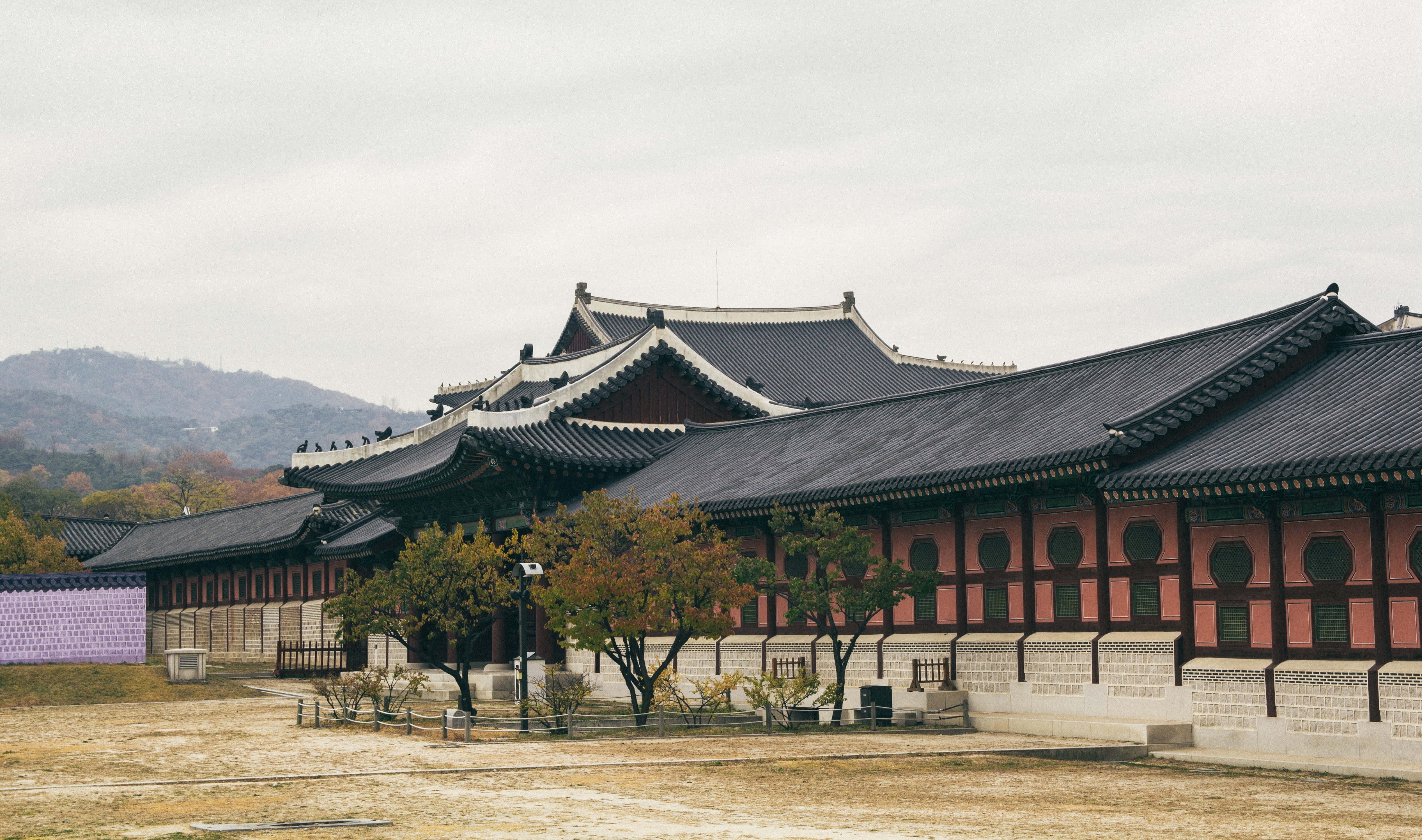Architecture 桌布 壁紙 — 傳統韓國宮殿建築，擁有彎曲的黑色屋頂和紅色牆壁，矗立在多雲的天空下，伴有秋樹和遠山。