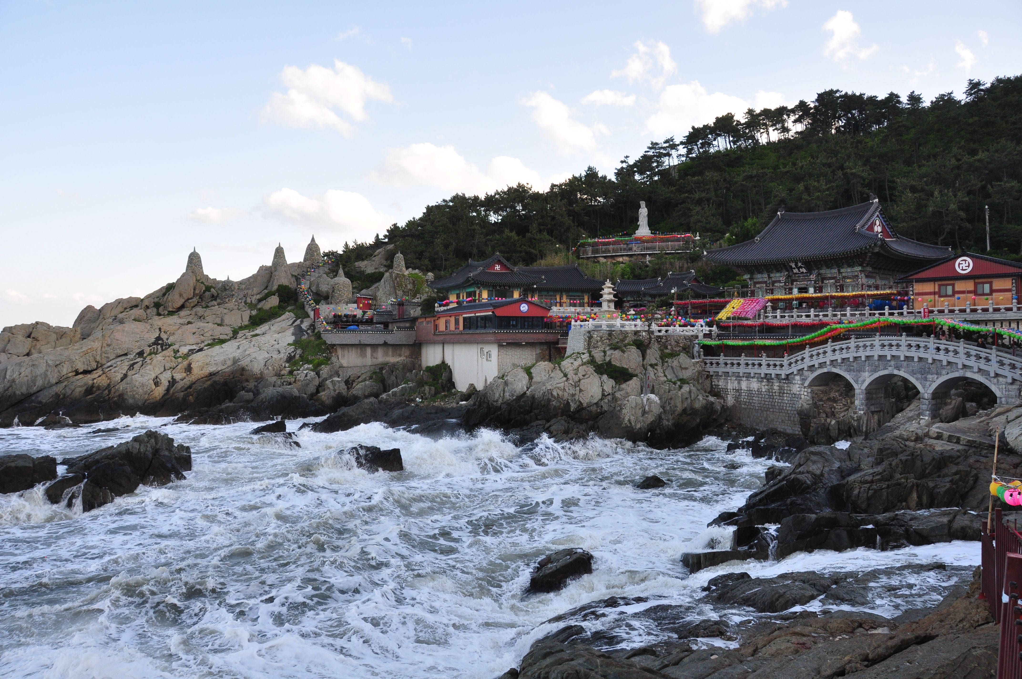 Busan 桌布 壁紙 — 傳統韓國寺廟坐落在岩石懸崖上，下方是波濤洶湧的海浪，天空部分多雲。