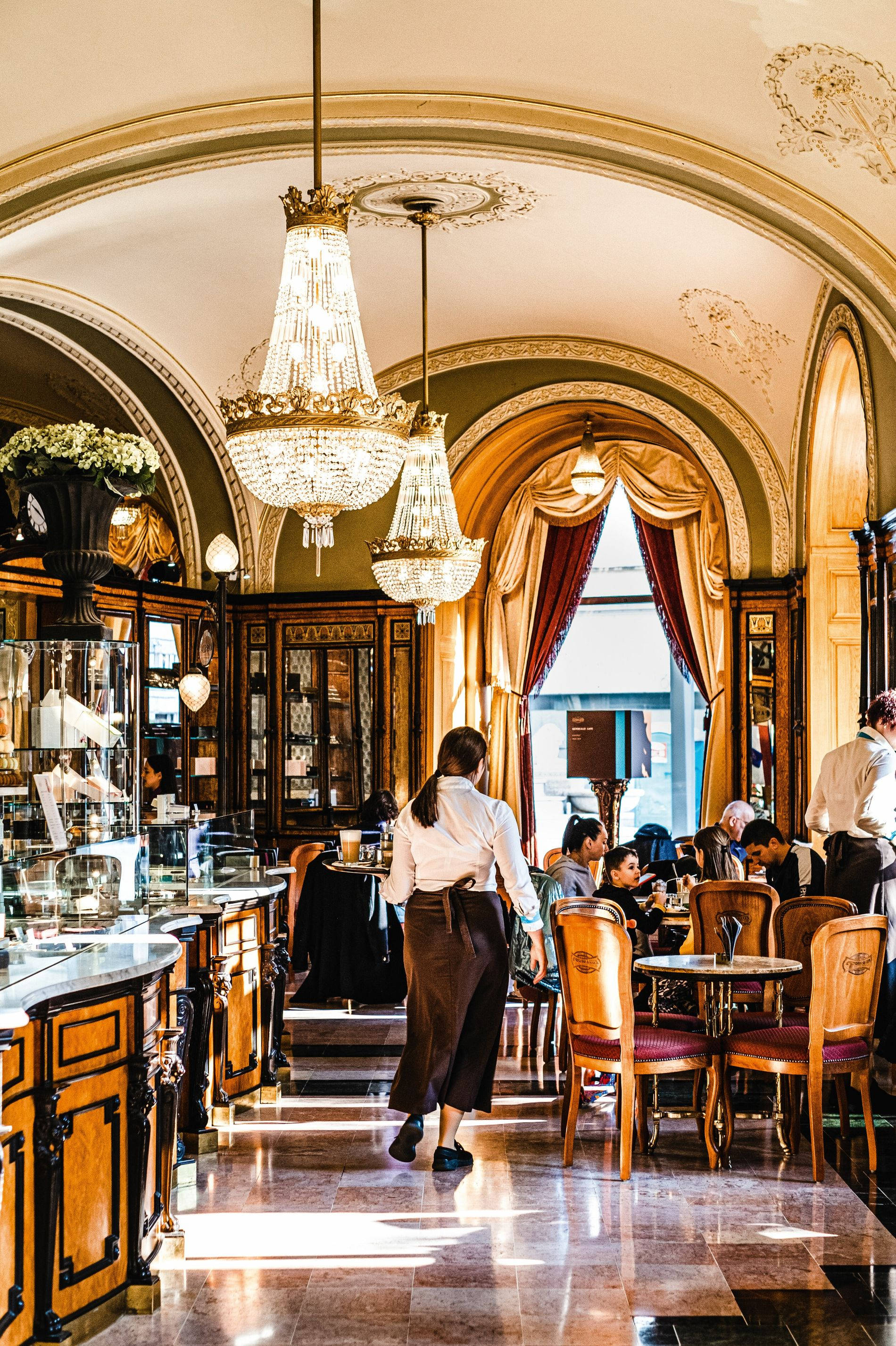 budapest 桌布 壁紙 — 優雅復古咖啡館，裝飾華麗的水晶吊燈、大理石地板，顧客在溫暖的金色燈光下享受下午茶。