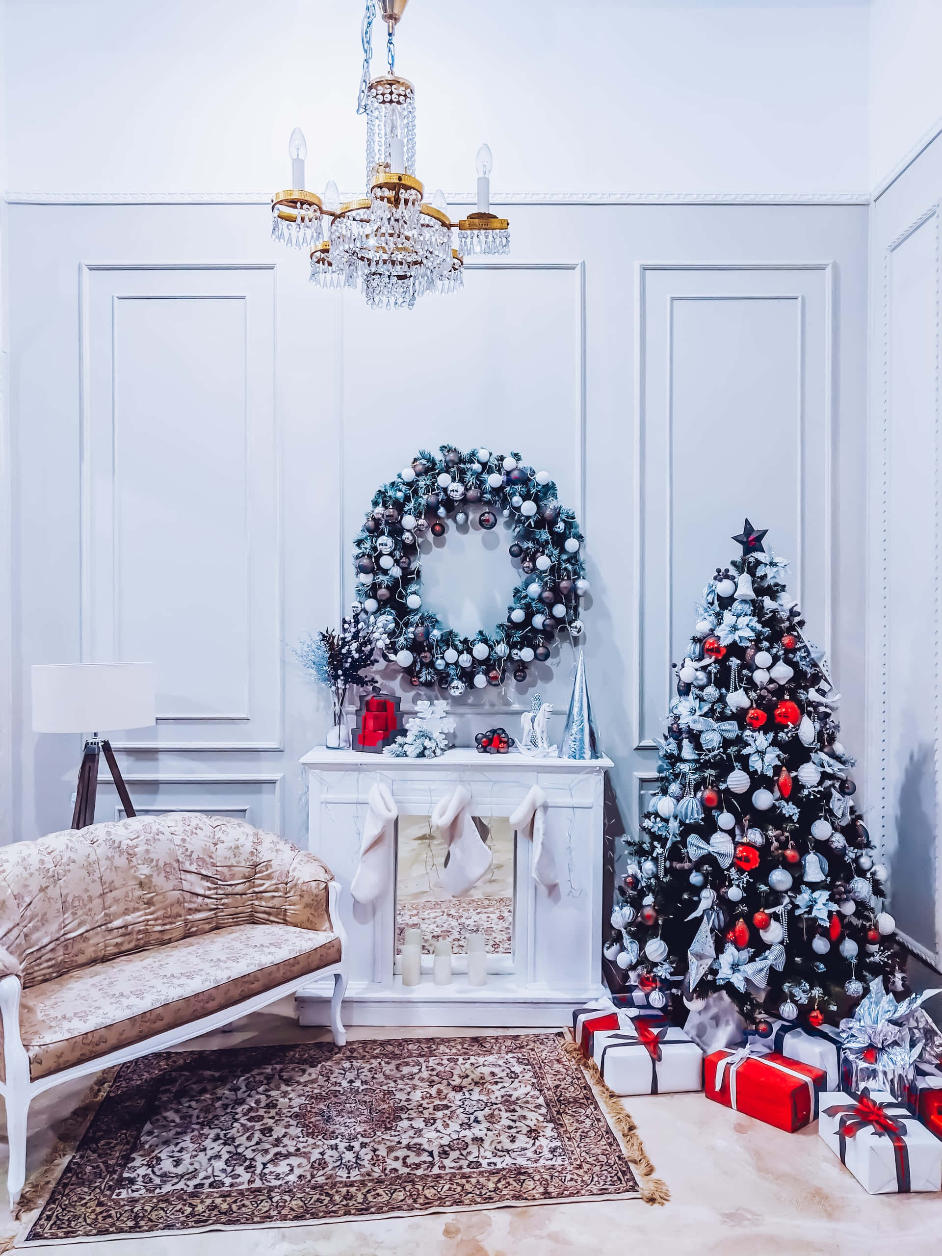 Aesthetic Christmas Tree 桌布 壁紙 — 優雅白色房間，聖誕裝飾有聖誕樹、紅色飾品、花環和水晶吊燈下的復古沙發。