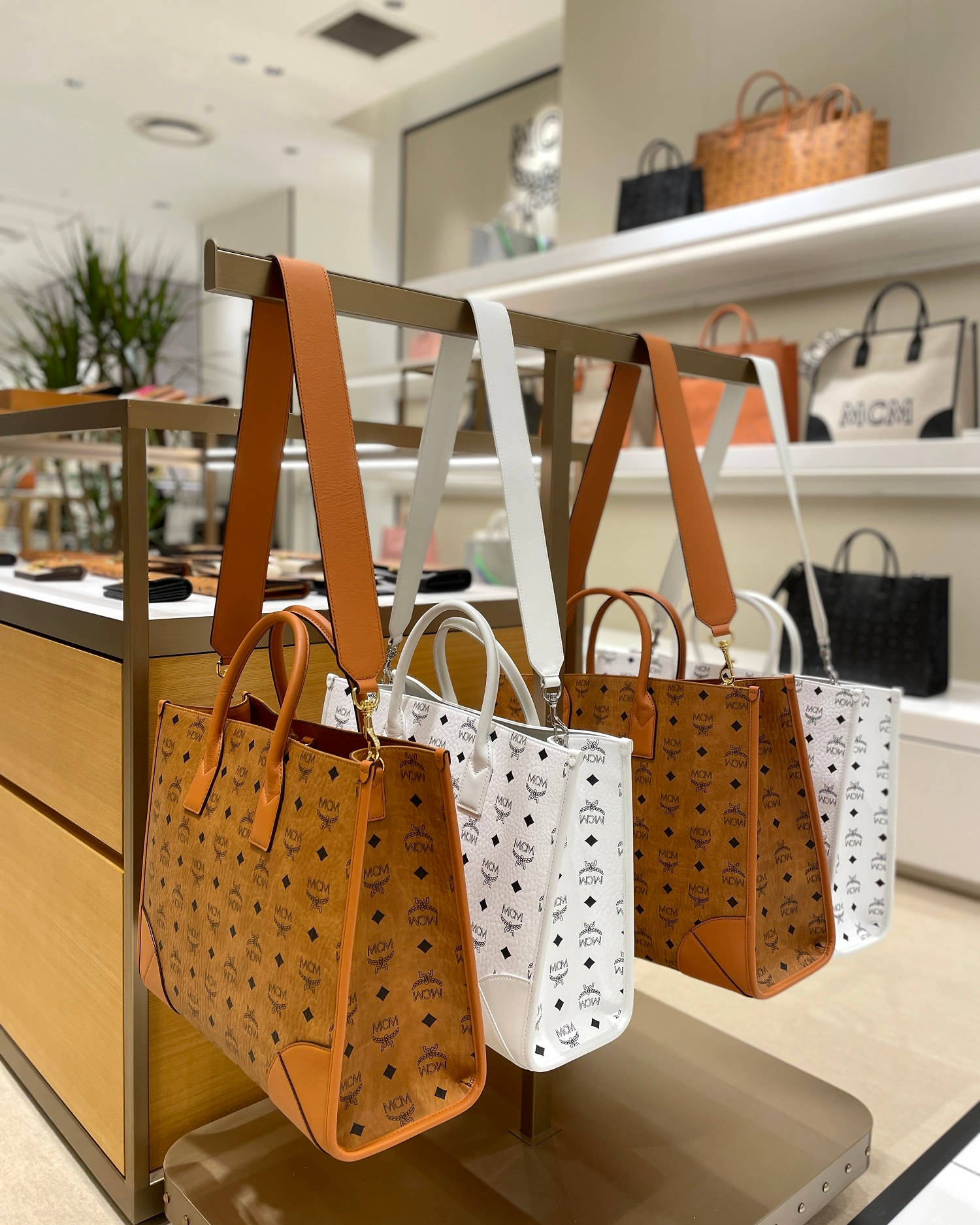 Store 桌布 壁紙 — 優雅的MCM手袋，採用棕褐色和白色，帶有標誌性字母組合圖案，整齊地懸掛在現代奢華零售精品店中。
