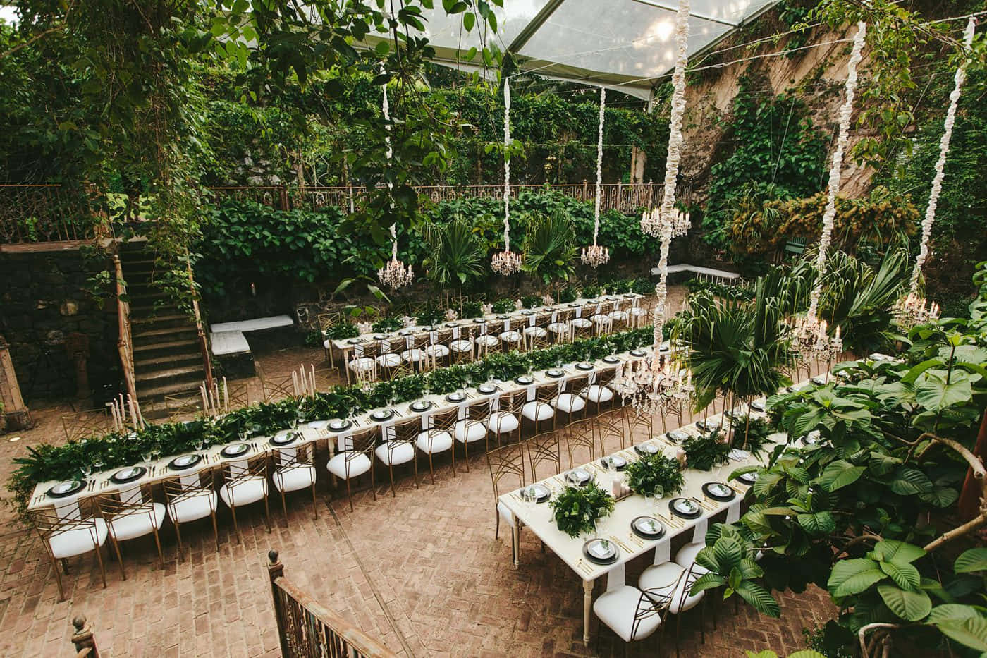 Wedding Aesthetic 桌布 壁紙 — 優雅的婚宴餐桌設置在玻璃屋頂下，周圍環繞著茂密的綠色植物和懸掛的水晶吊燈。