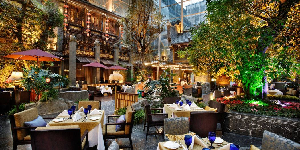 Chengdu 桌布 壁紙 — 優雅的室內庭院餐廳，在溫暖的燈籠下設有餐桌，周圍環繞著茂密的綠色植物和石頭建築。