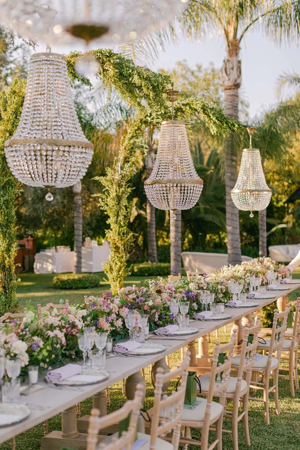 Wedding Aesthetic 桌布 壁紙 — 優雅的戶外婚宴餐桌，裝飾有花卉佈置、水晶吊燈和木椅，位於棕櫚樹下。